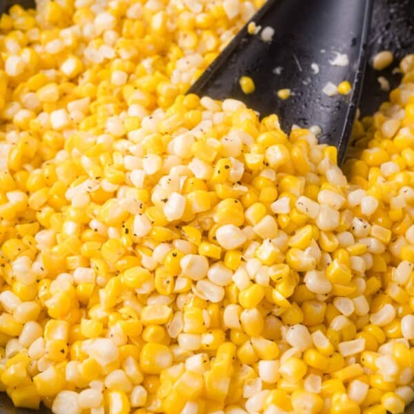 Close-up of cooked yellow and white corn kernels with black specks of seasoning, being stirred with a black spatula.