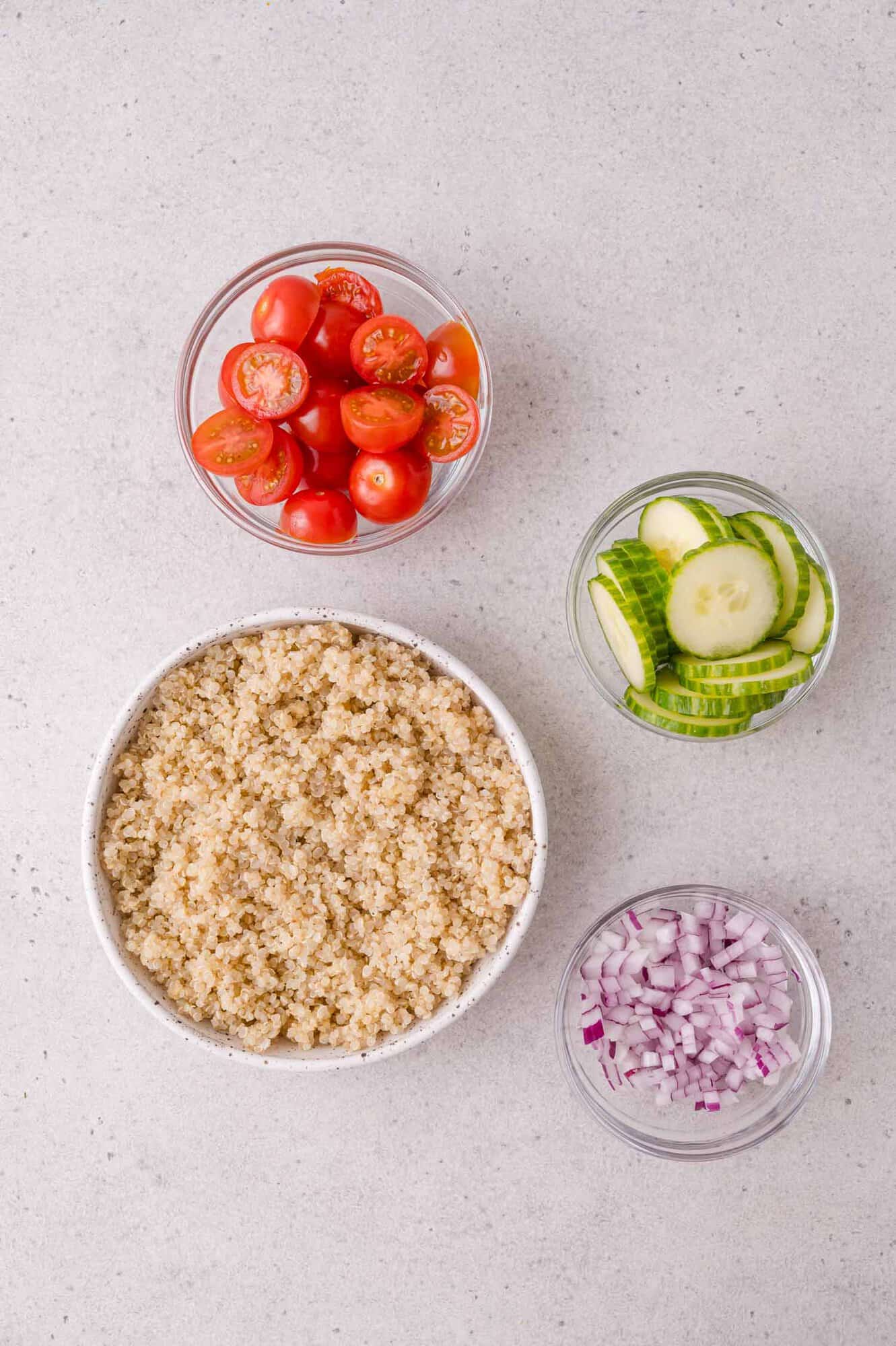A bowl of cooked quinoa, and three smaller bowls containing halved cherry tomatoes, sliced cucumbers, and diced red onions, all on a light countertop.