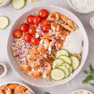 Bowl with quinoa, grilled salmon, cherry tomatoes, cucumbers, diced red onion, feta cheese, and tzatziki sauce.