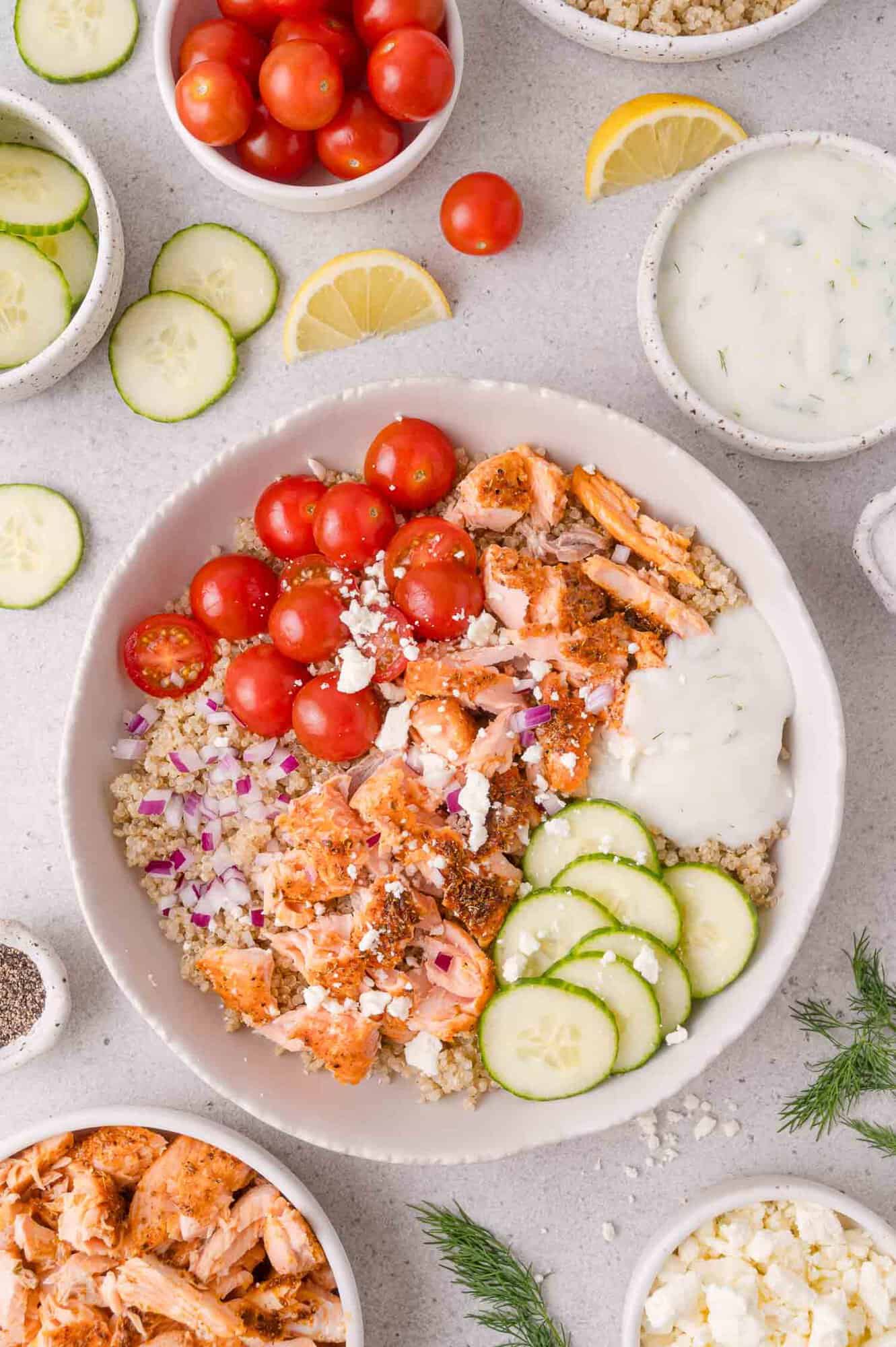 Bowl with quinoa, cooked salmon, cherry tomatoes, cucumber slices, red onion, feta, and tzatziki sauce. Surrounded by small bowls of ingredients and lemon wedges on a white background.
