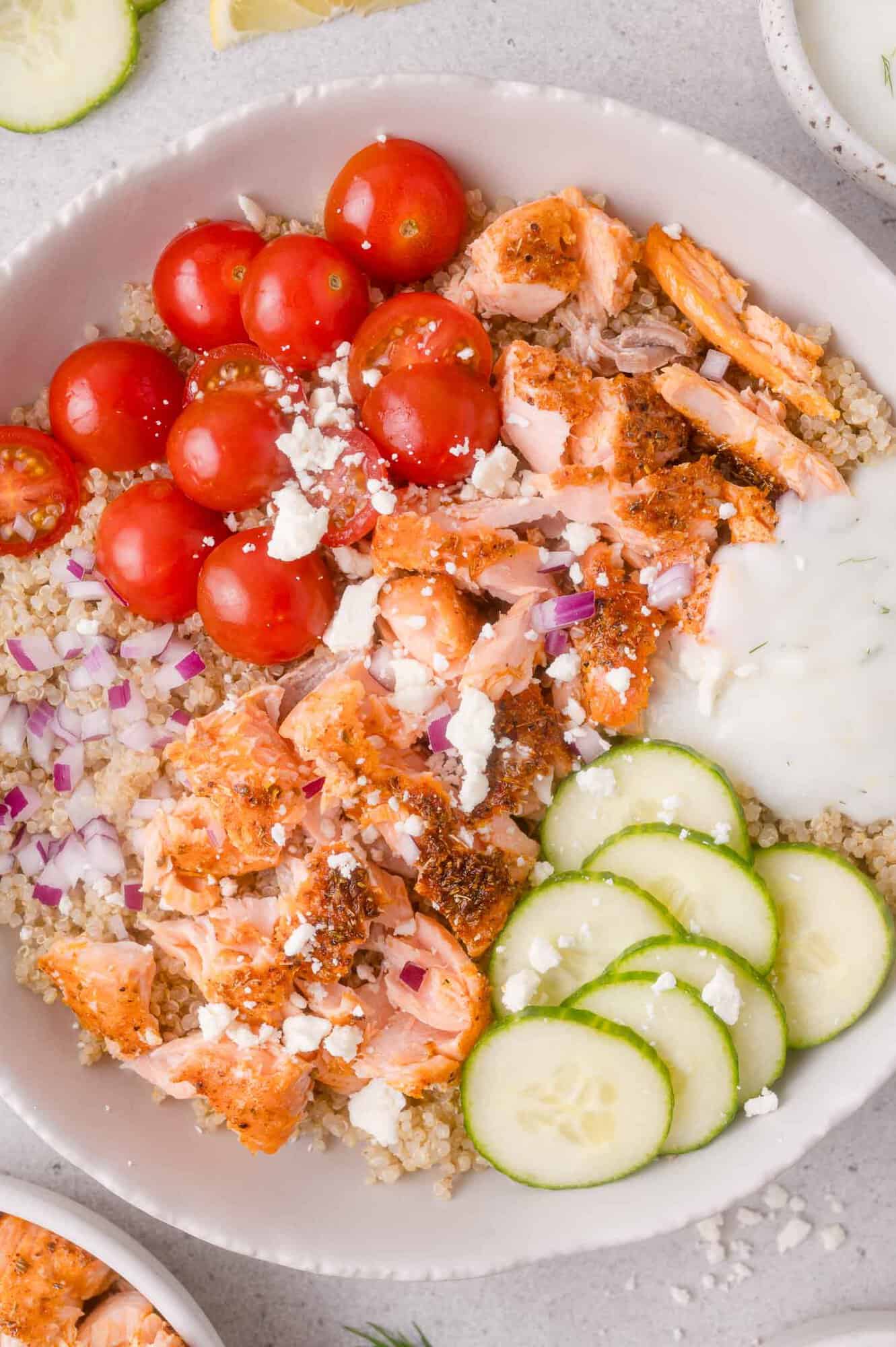 A bowl with quinoa, flaked salmon, cherry tomatoes, cucumber slices, red onion, crumbled feta, and a dollop of white sauce.