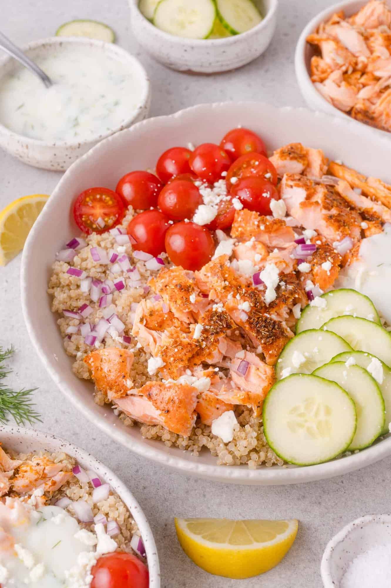 Bowl with quinoa, sliced cucumbers, cherry tomatoes, red onions, crumbled feta, and seasoned salmon, served with a side of creamy dressing and lemon wedges.