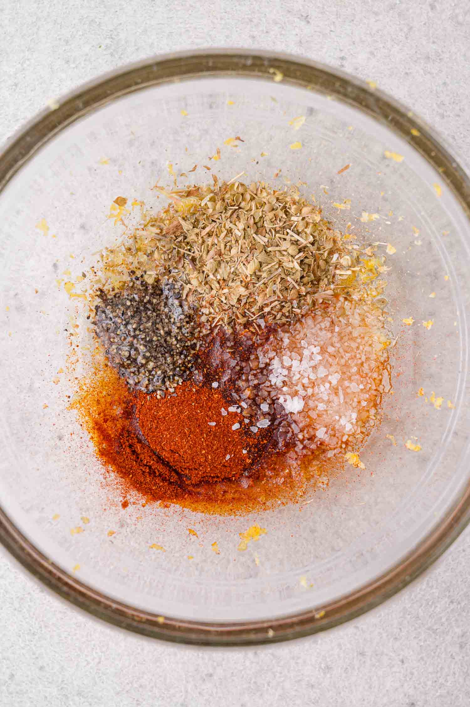 A glass bowl containing piles of dried oregano, ground black pepper, salt, paprika, and lemon zest on a light surface.