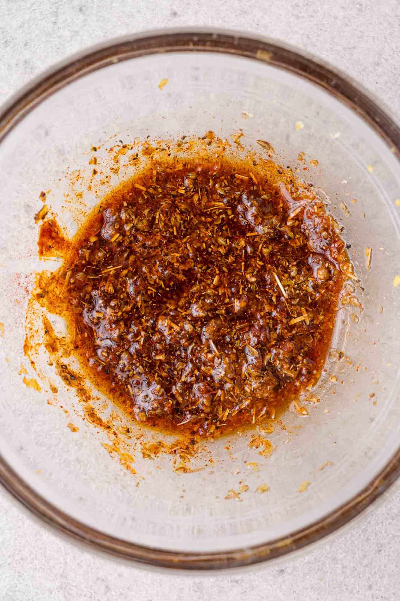A glass bowl containing a reddish-brown spice marinade with visible herbs and oil, placed on a light surface.