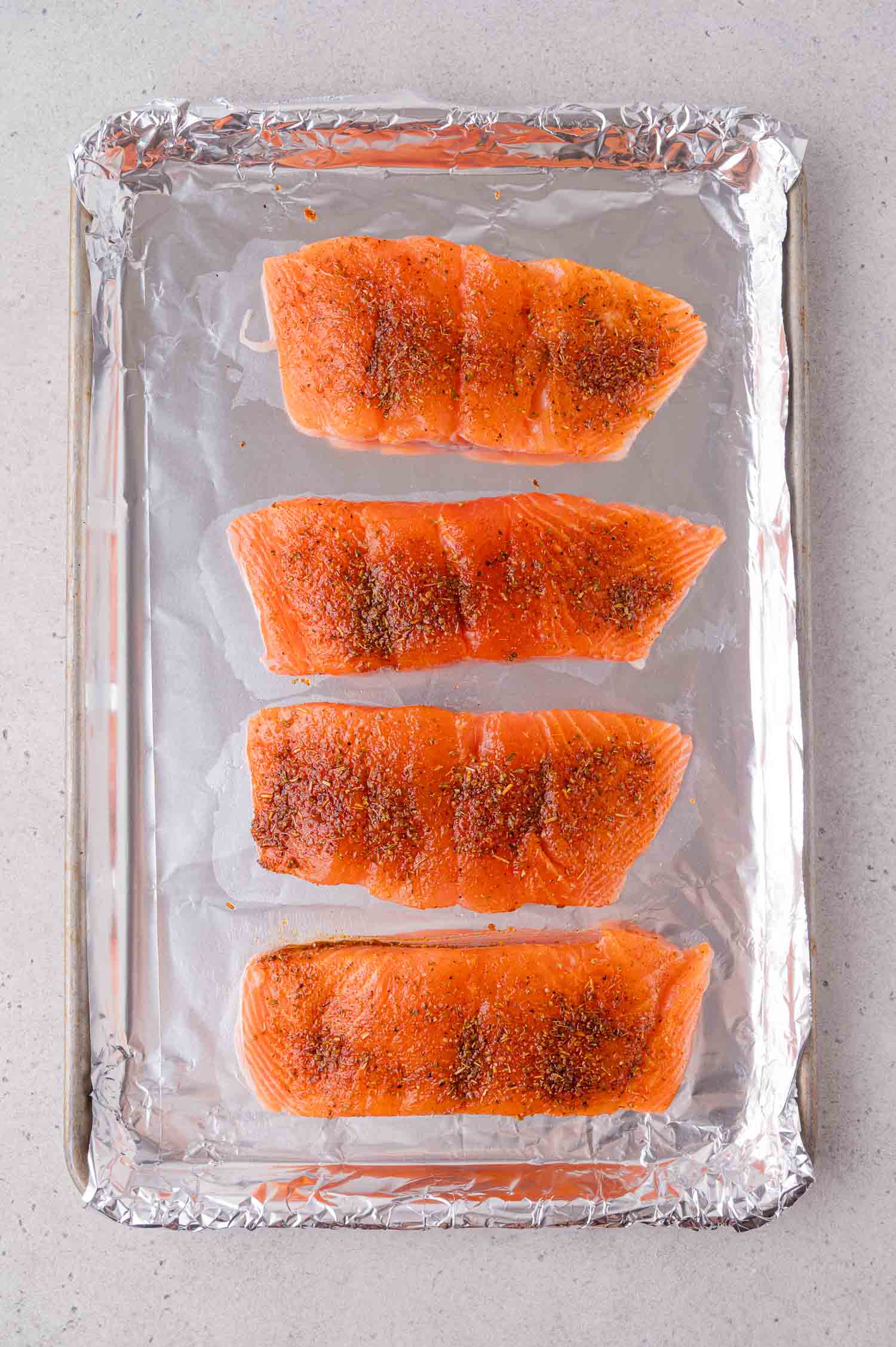 Four raw salmon fillets with seasoning are arranged in a row on a foil-lined baking sheet.