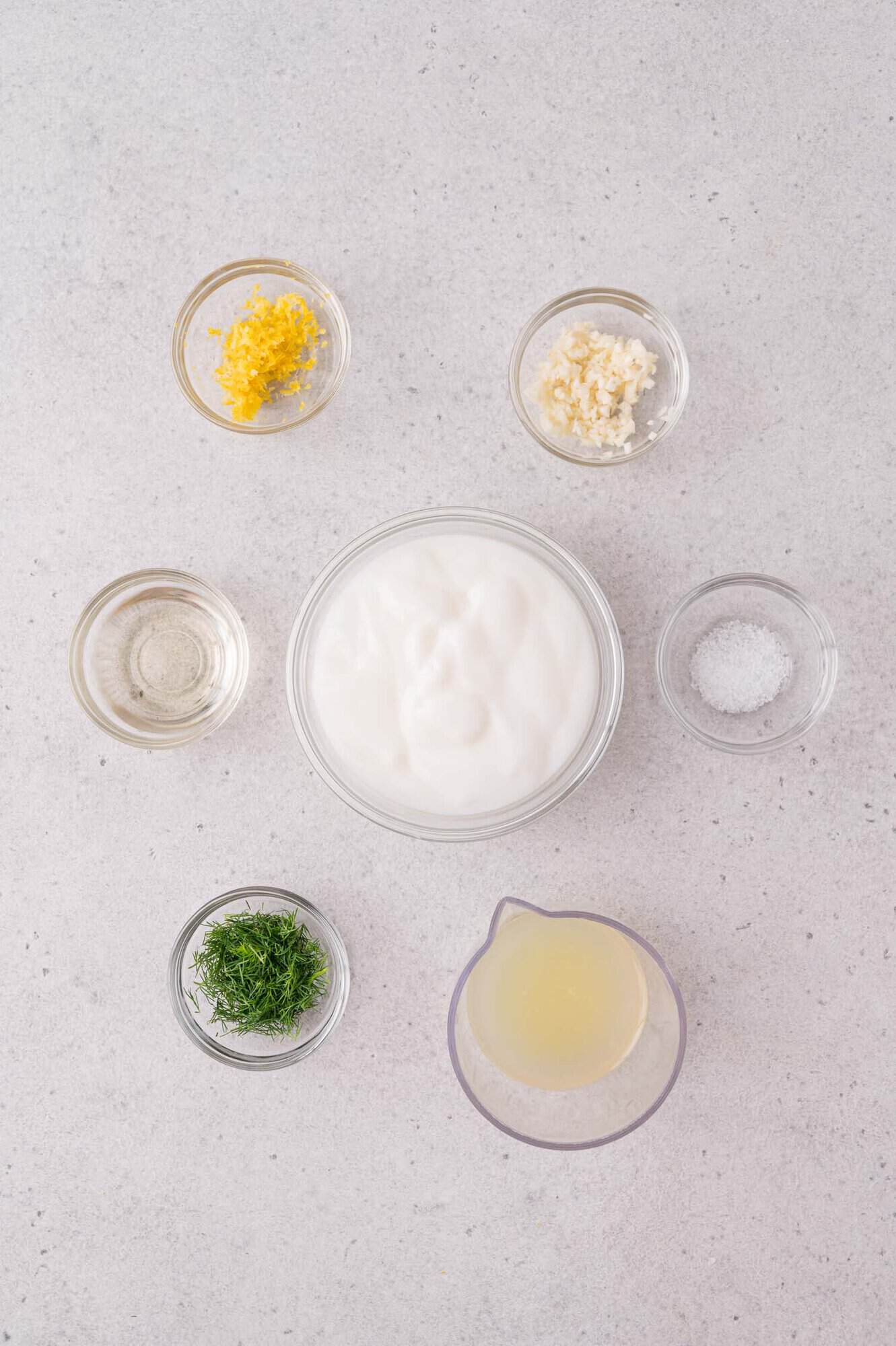 Six small bowls containing yogurt, chopped dill, minced garlic, lemon zest, salt, lemon juice, and vinegar arranged on a light-colored surface.