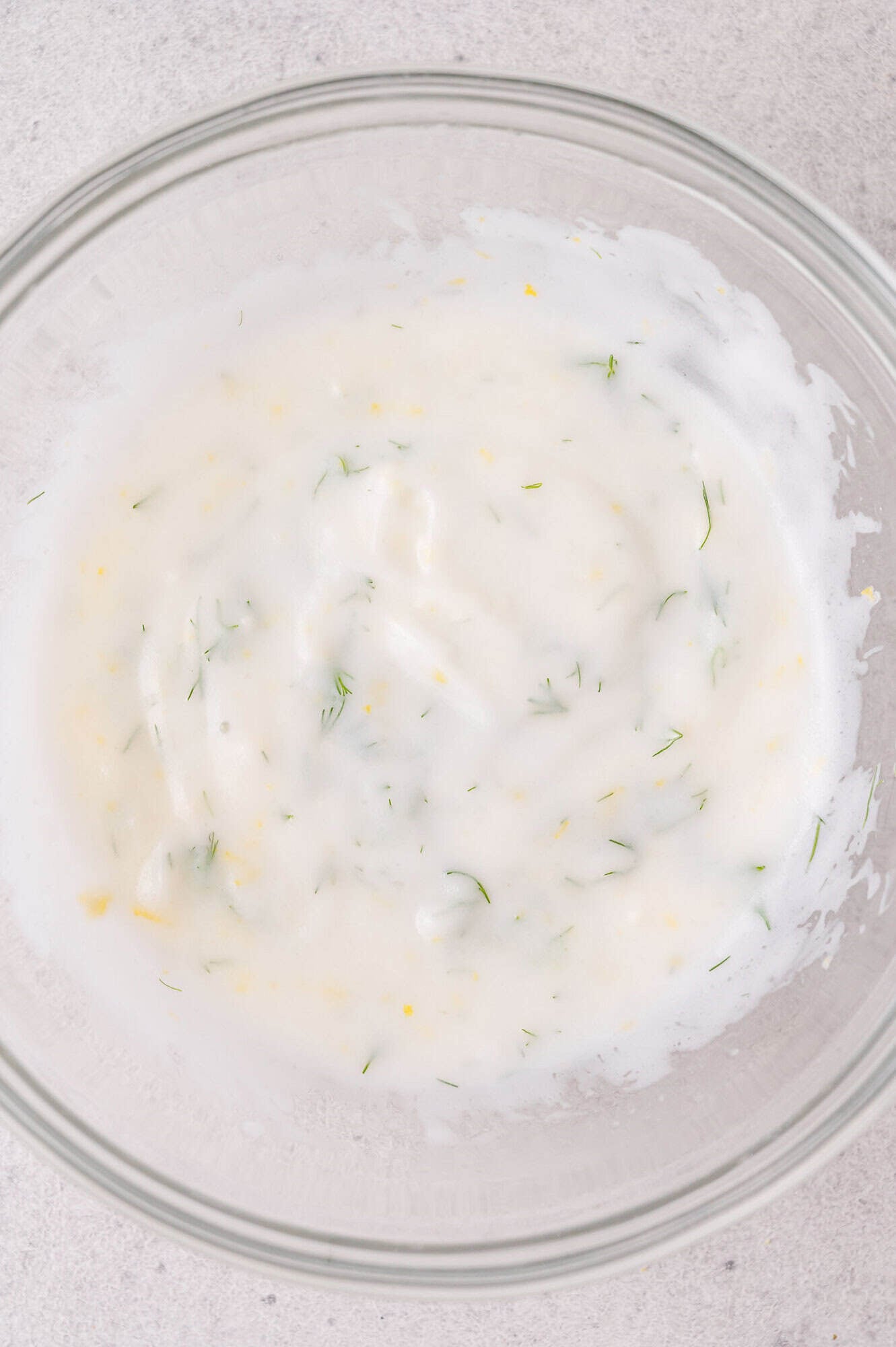 Clear glass bowl containing a white, creamy sauce with dill mixed in, set on a light-colored surface.