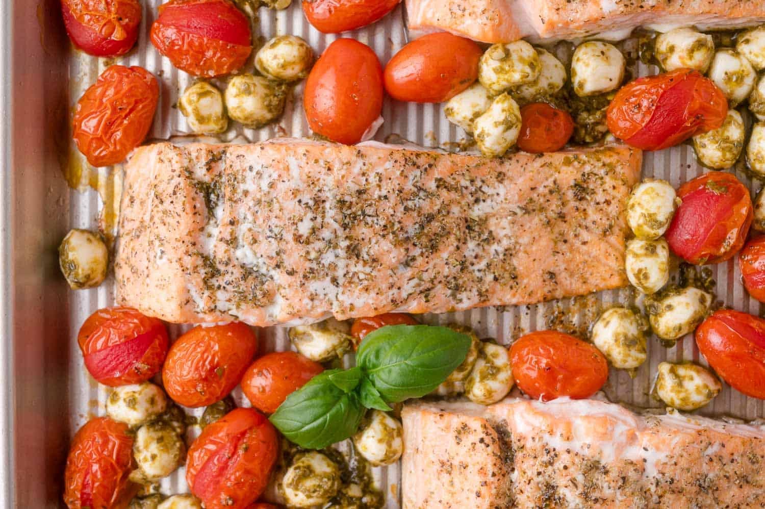 Baked salmon fillets with cherry tomatoes and mozzarella balls, garnished with pesto and fresh basil, on a metal baking sheet.