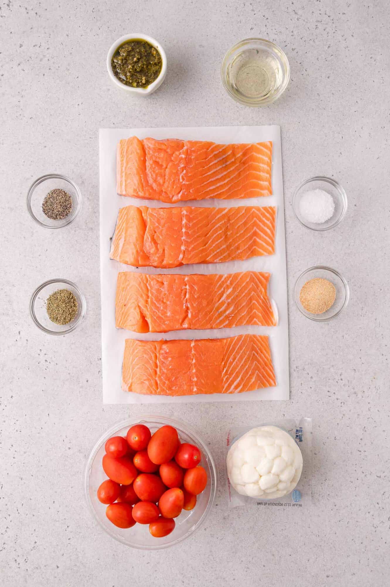 Four raw salmon fillets on parchment paper, surrounded by bowls of grape tomatoes, mozzarella, seasonings, oil, and pesto on a gray countertop.
