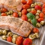 Baked salmon fillets on a sheet pan with roasted cherry tomatoes, mozzarella balls with pesto, and fresh basil leaves.