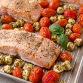 Baked salmon fillets on a sheet pan with roasted cherry tomatoes, mozzarella balls with pesto, and fresh basil leaves.