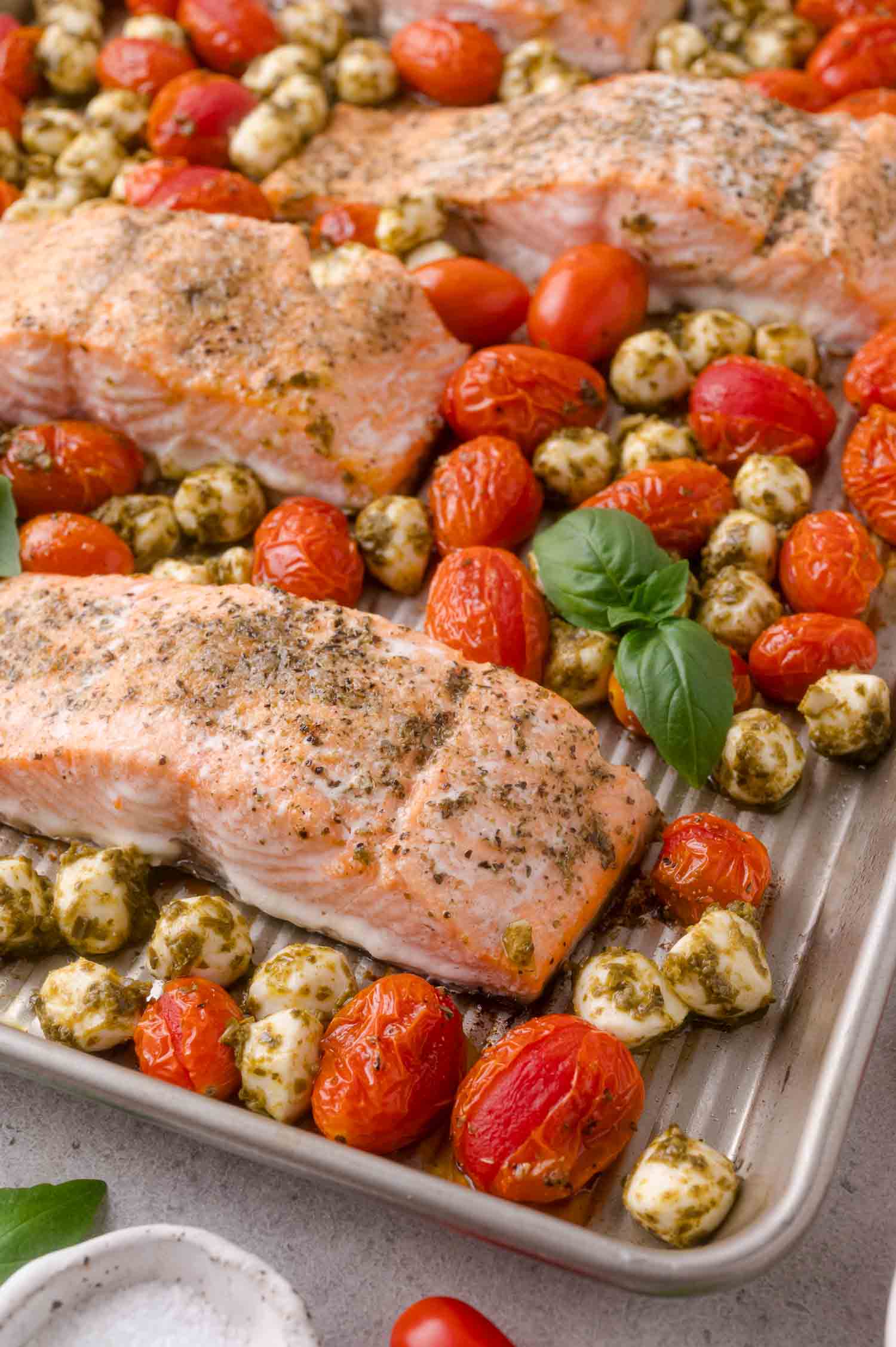 Baked salmon fillets on a sheet pan with roasted grape tomatoes, pesto-coated mozzarella balls, and fresh basil leaves.