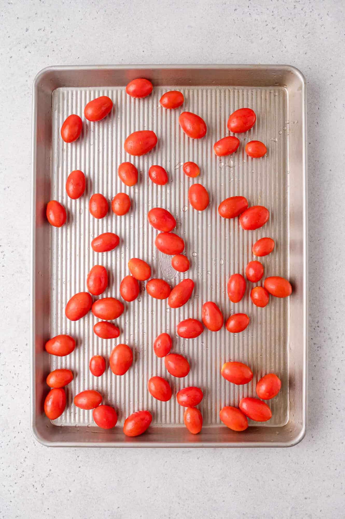 A baking sheet with raw grape tomatoes spread out evenly on a light-colored countertop.