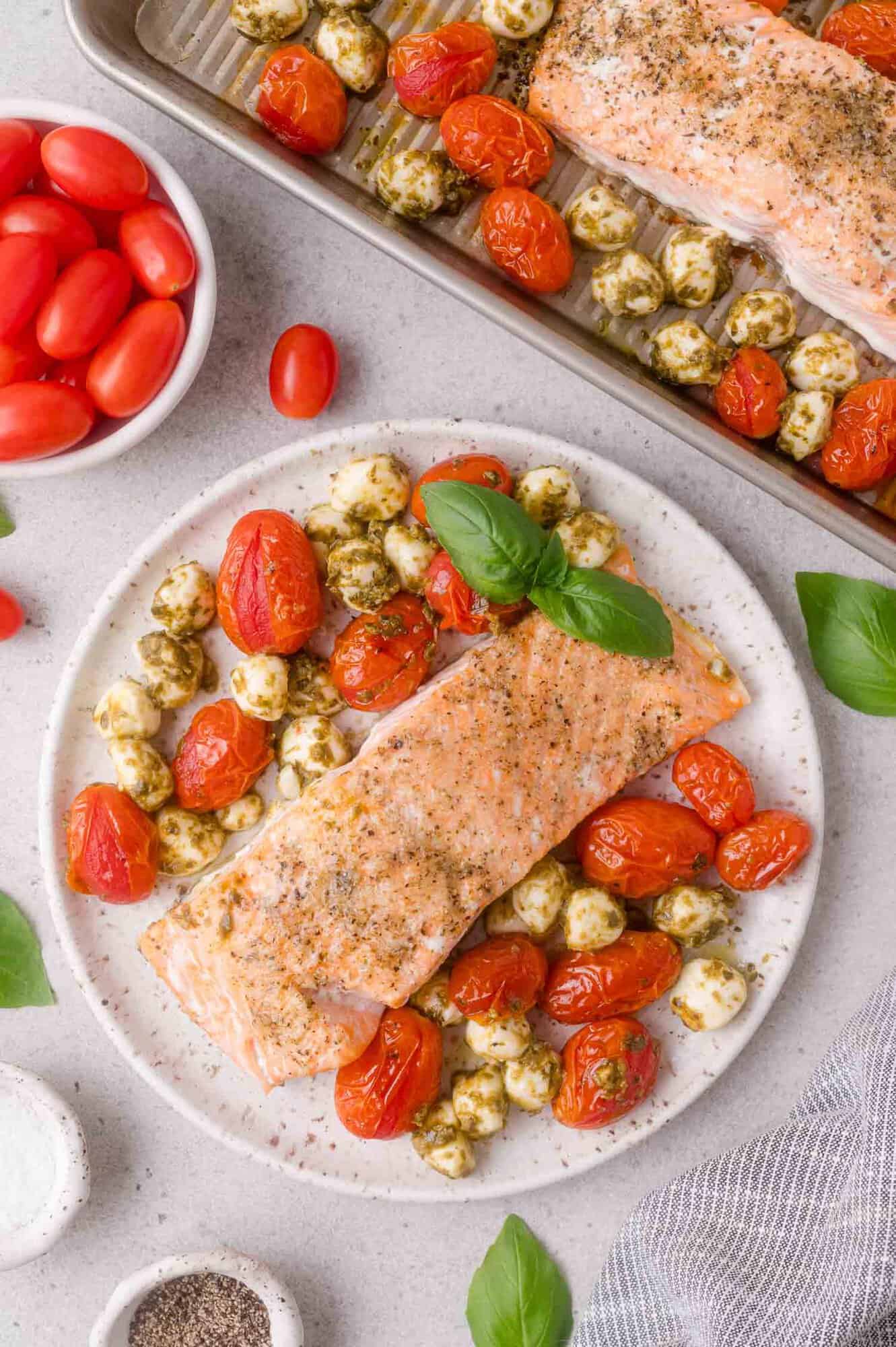 Baked salmon fillet with roasted cherry tomatoes and mozzarella balls garnished with basil, served on a white plate next to a tray with more salmon and tomatoes.
