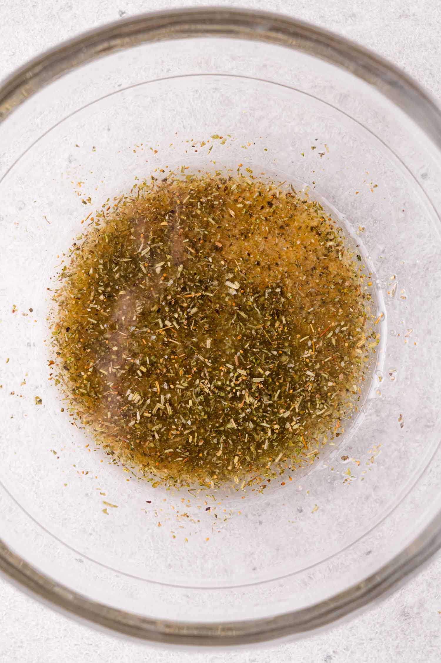 Clear glass bowl containing a mixture of oil and dried herbs on a light surface.