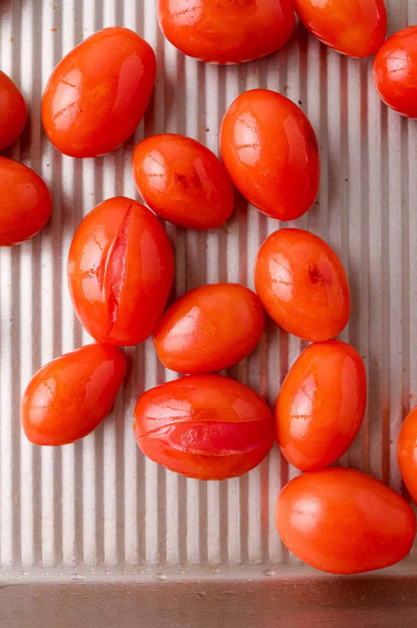 Several oval-shaped, bright red tomatoes are arranged on a ridged metal baking tray, some with splits in their skins.