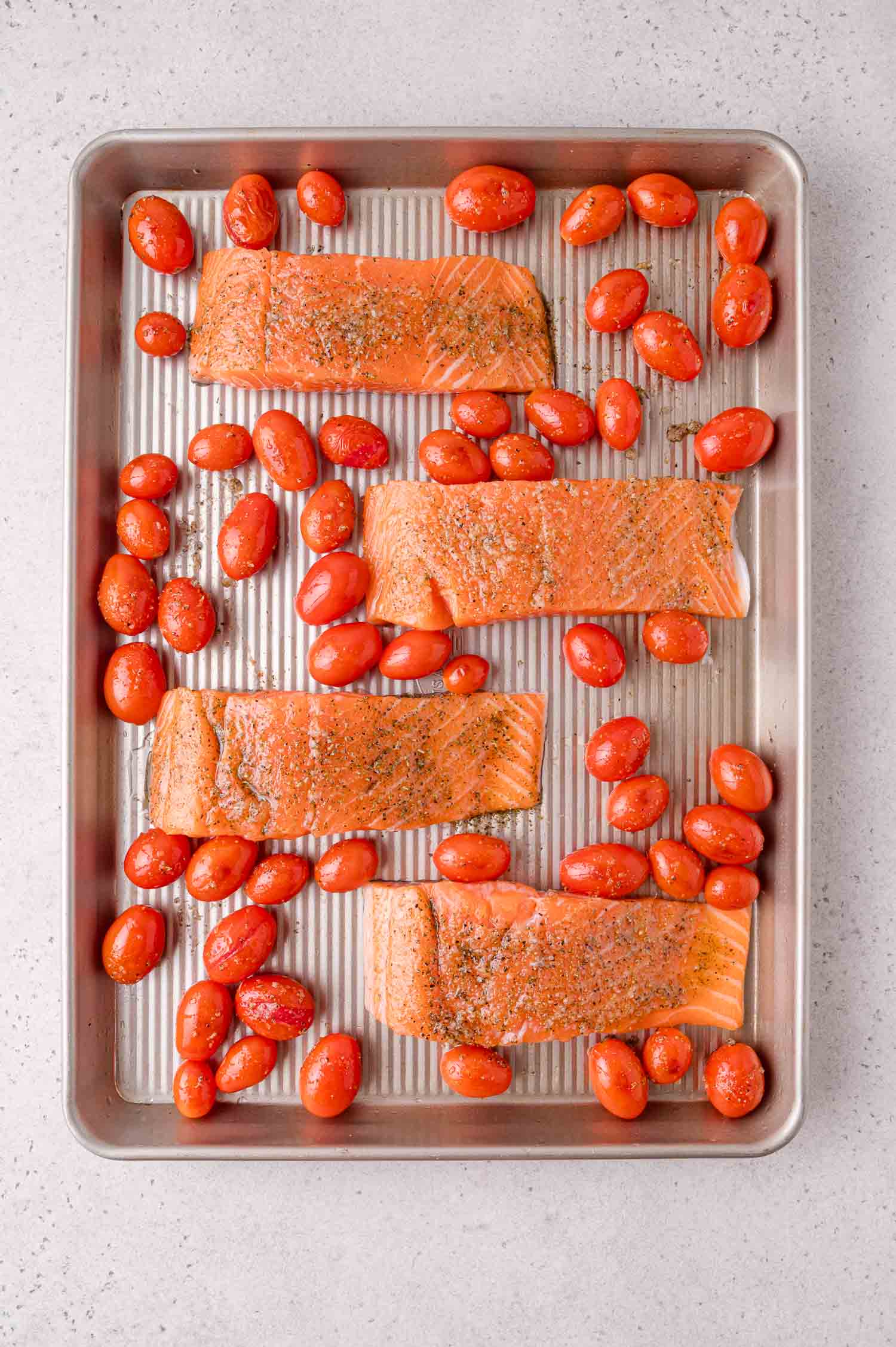 Four raw salmon fillets and grape tomatoes are arranged on a metal baking sheet, seasoned with herbs and pepper, ready for roasting.