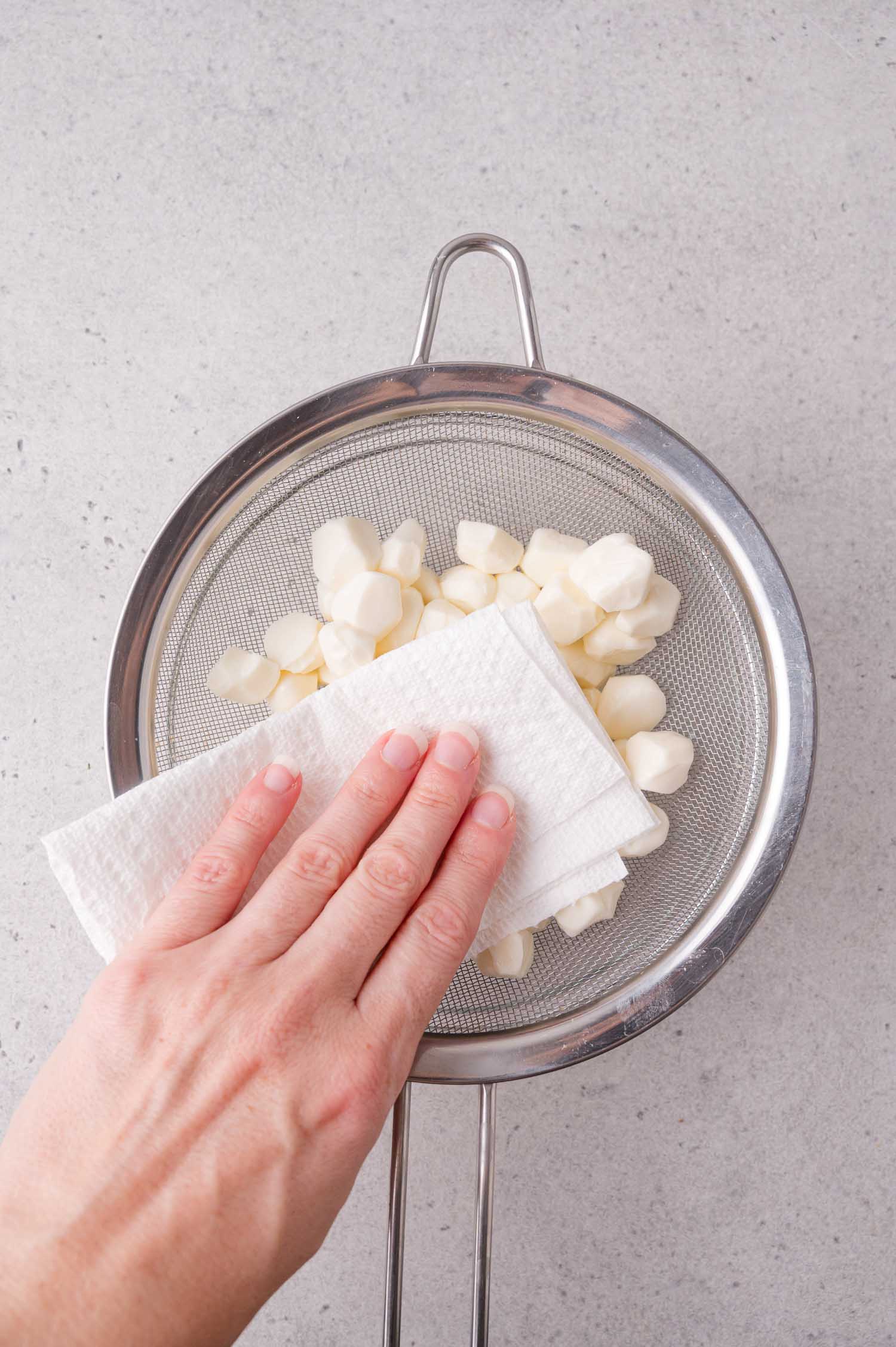 A hand presses a paper towel on mini mozzarella balls in a metal strainer, on a light gray countertop.