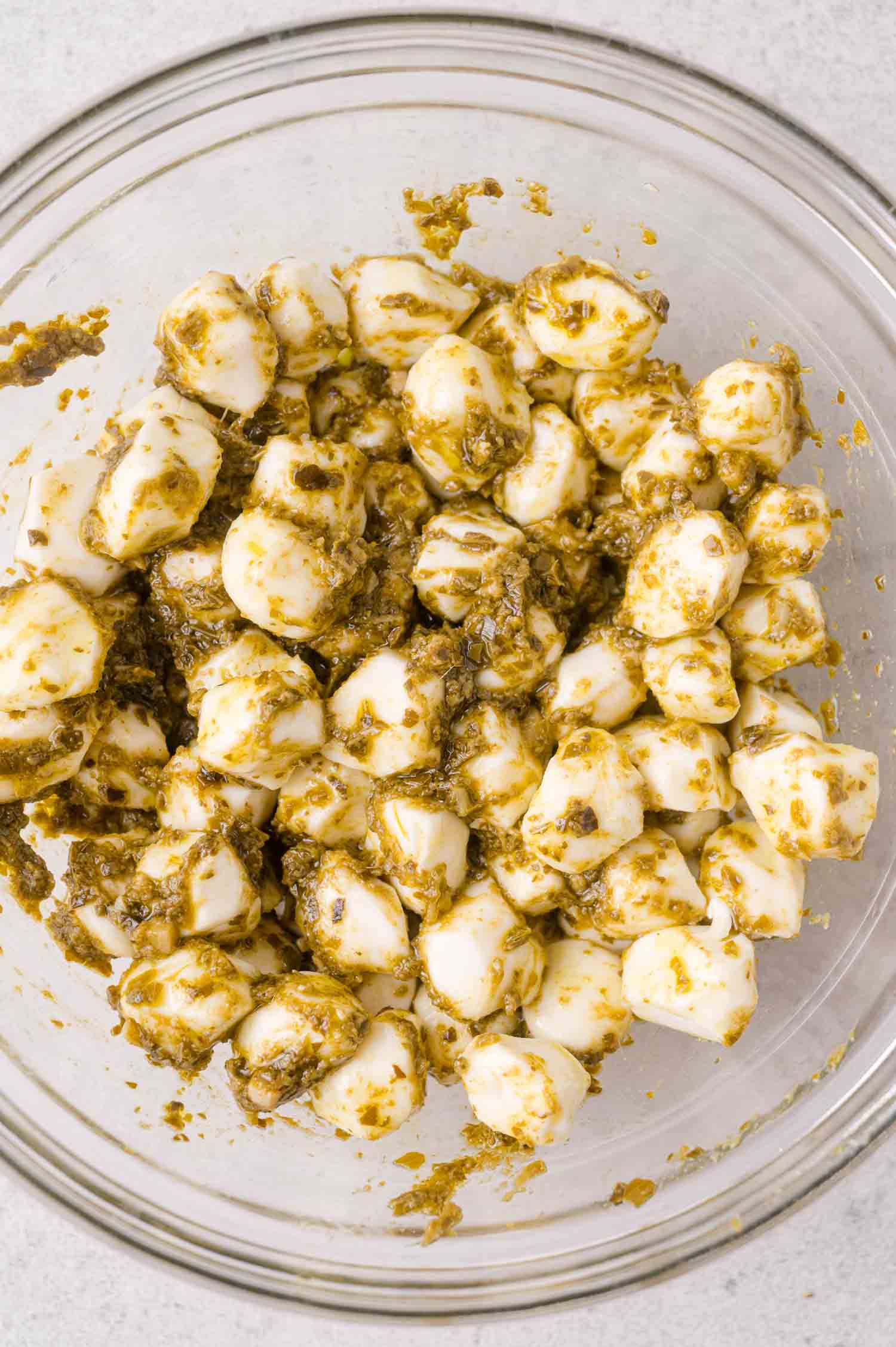 A glass bowl filled with small mozzarella balls coated in green pesto sauce, placed on a light-colored surface.
