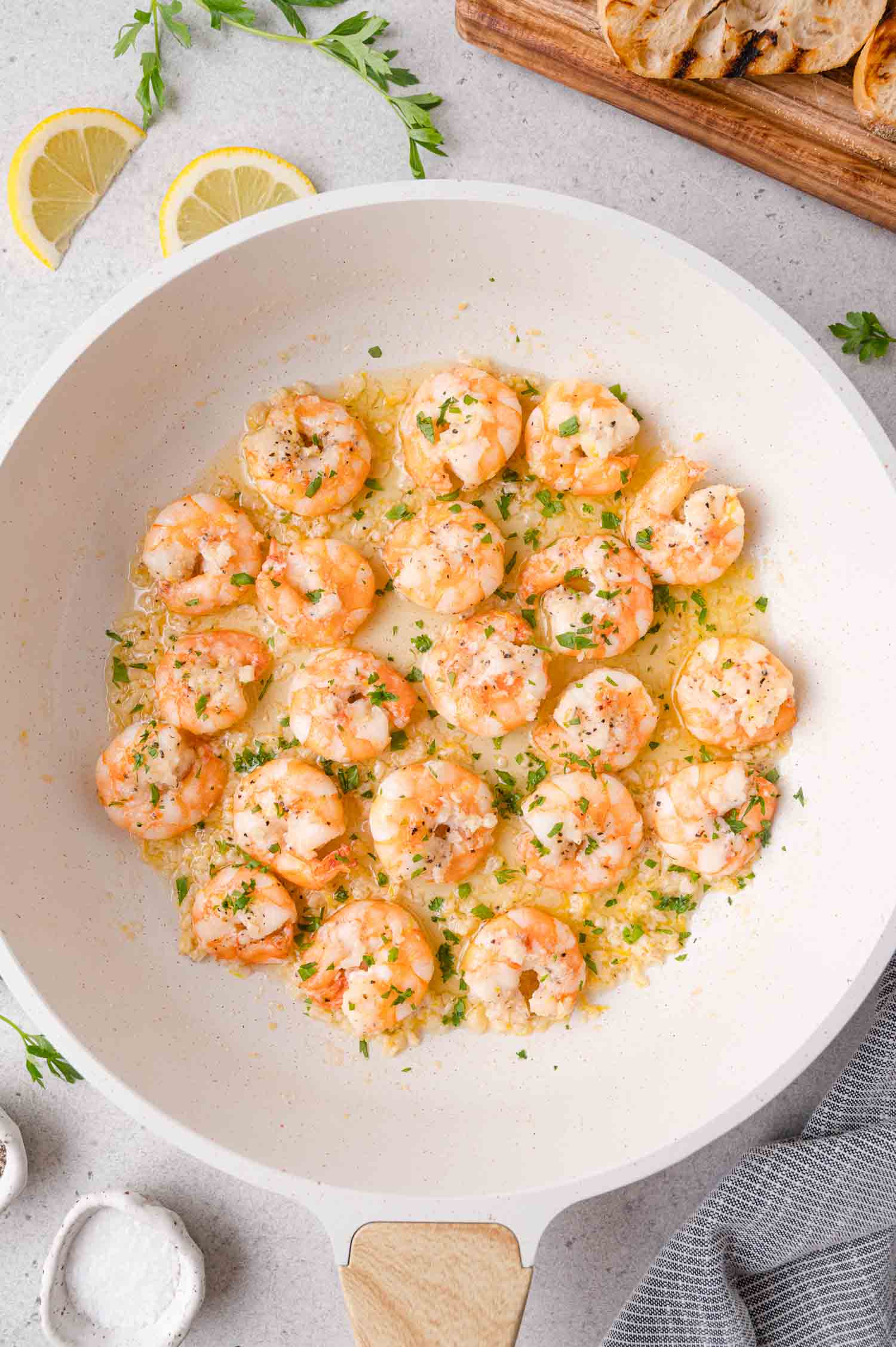 Shrimp scampi cooked in a white skillet with garlic, herbs, and lemon butter sauce, garnished with parsley. Lemon slices and grilled bread are visible in the background.