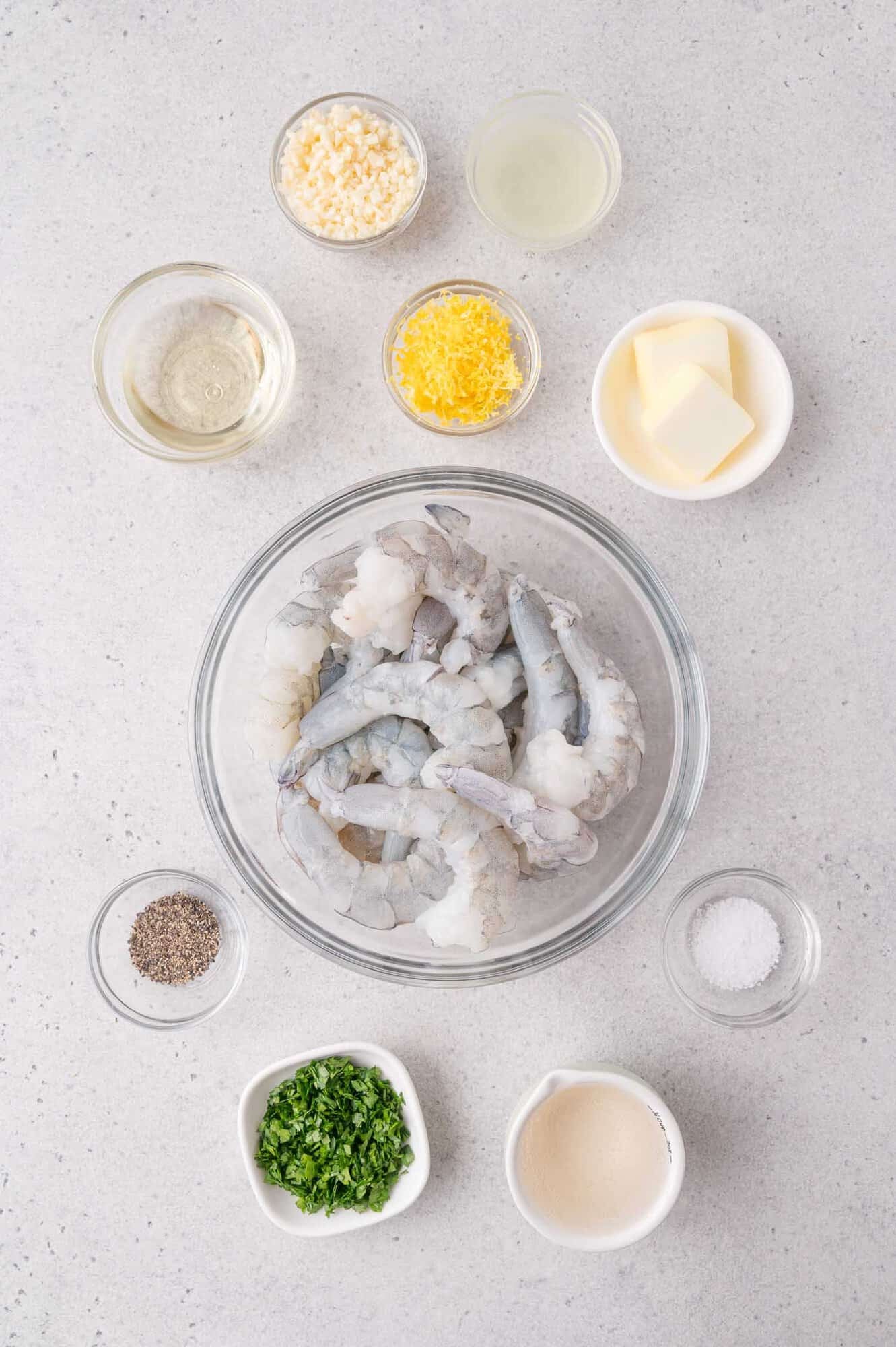 Raw shrimp in a glass bowl surrounded by bowls of chopped garlic, lemon zest, lemon juice, butter, salt, pepper, parsley, white wine, and cream on a light countertop.
