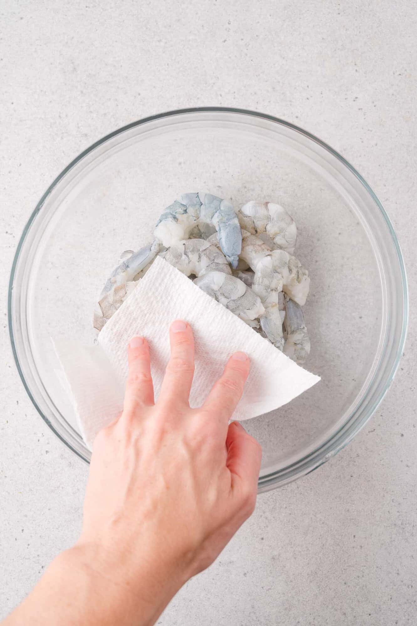 A hand uses a paper towel to pat dry raw, peeled shrimp in a clear glass bowl on a light-colored surface.