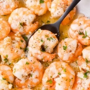 Cooked shrimp topped with minced garlic and chopped herbs, served in a garlic butter sauce with a black spoon scooping a piece.