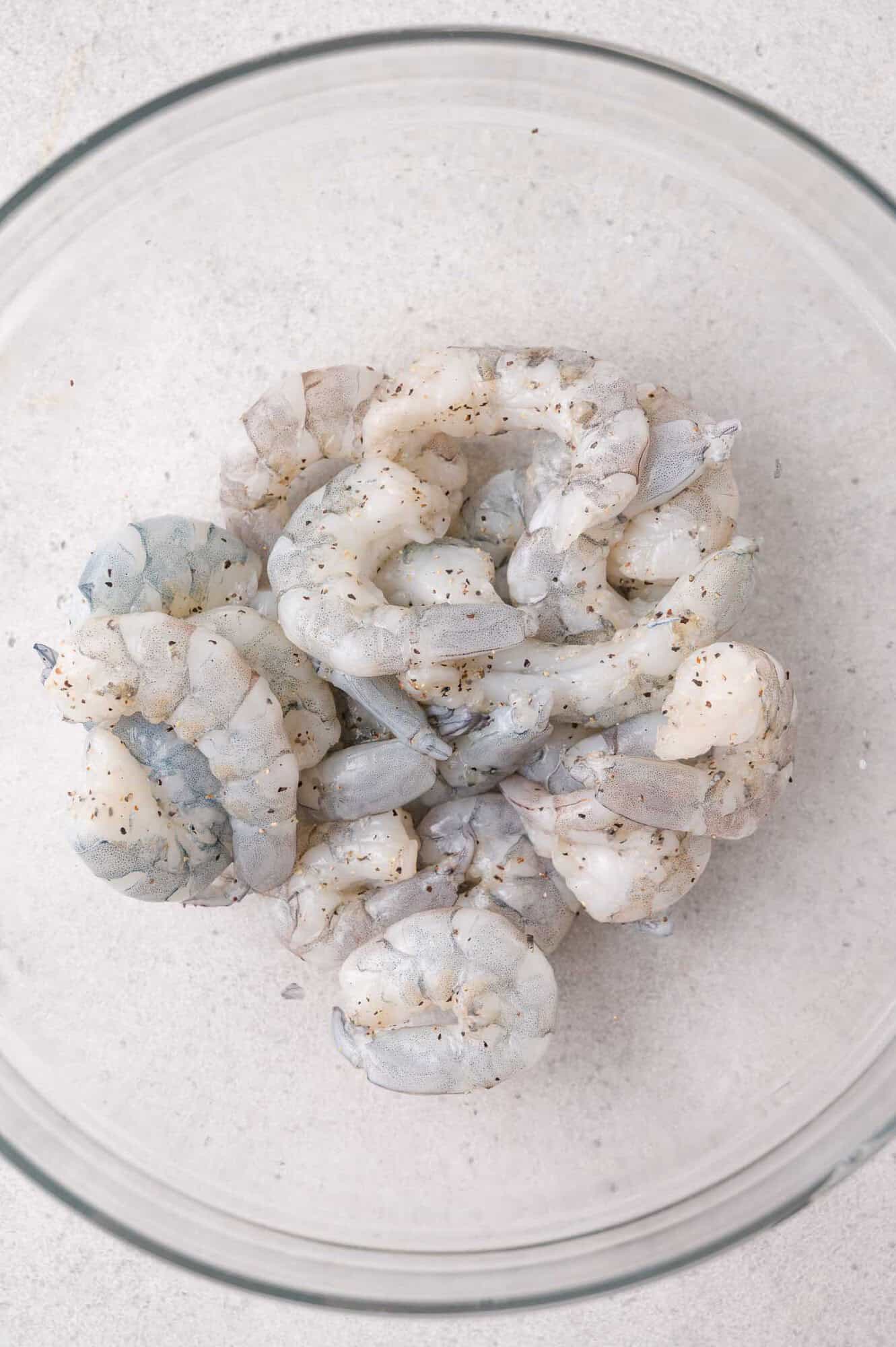 Raw, peeled shrimp seasoned with ground black pepper in a clear glass bowl on a light-colored surface.