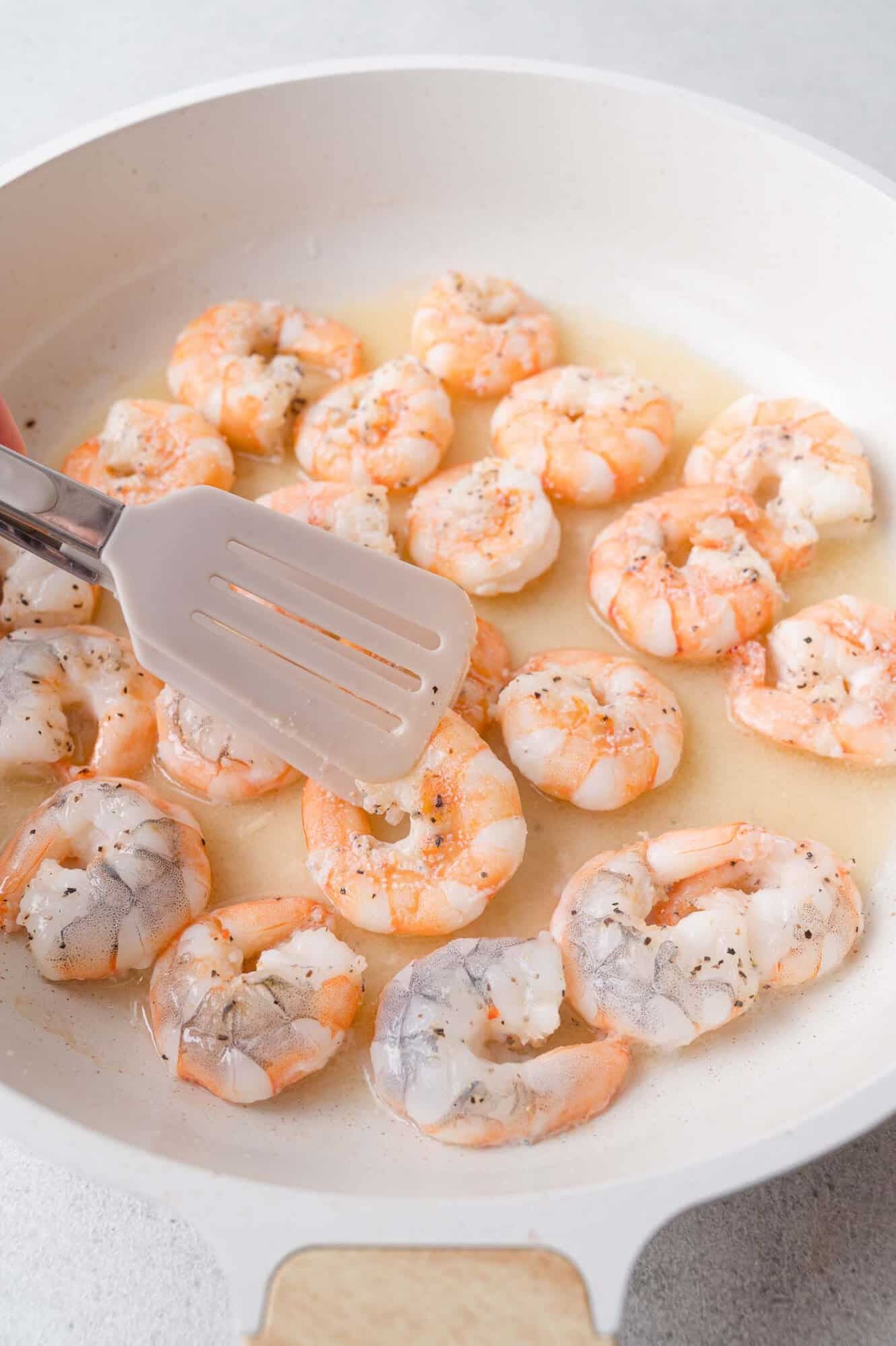 A spatula holds a cooked shrimp above a white skillet filled with shrimp simmering in a light sauce.