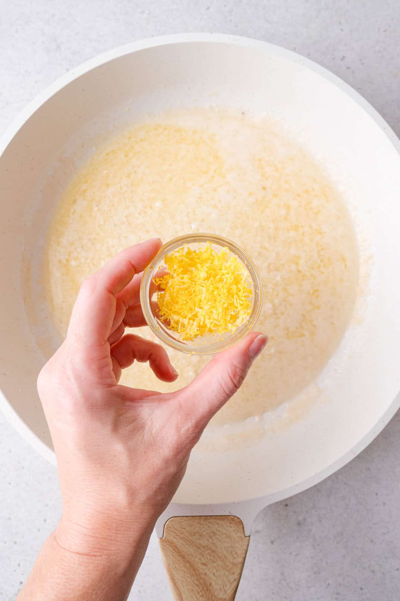 A hand holds a small bowl of lemon zest above a white pan containing melted butter on a light-colored surface.