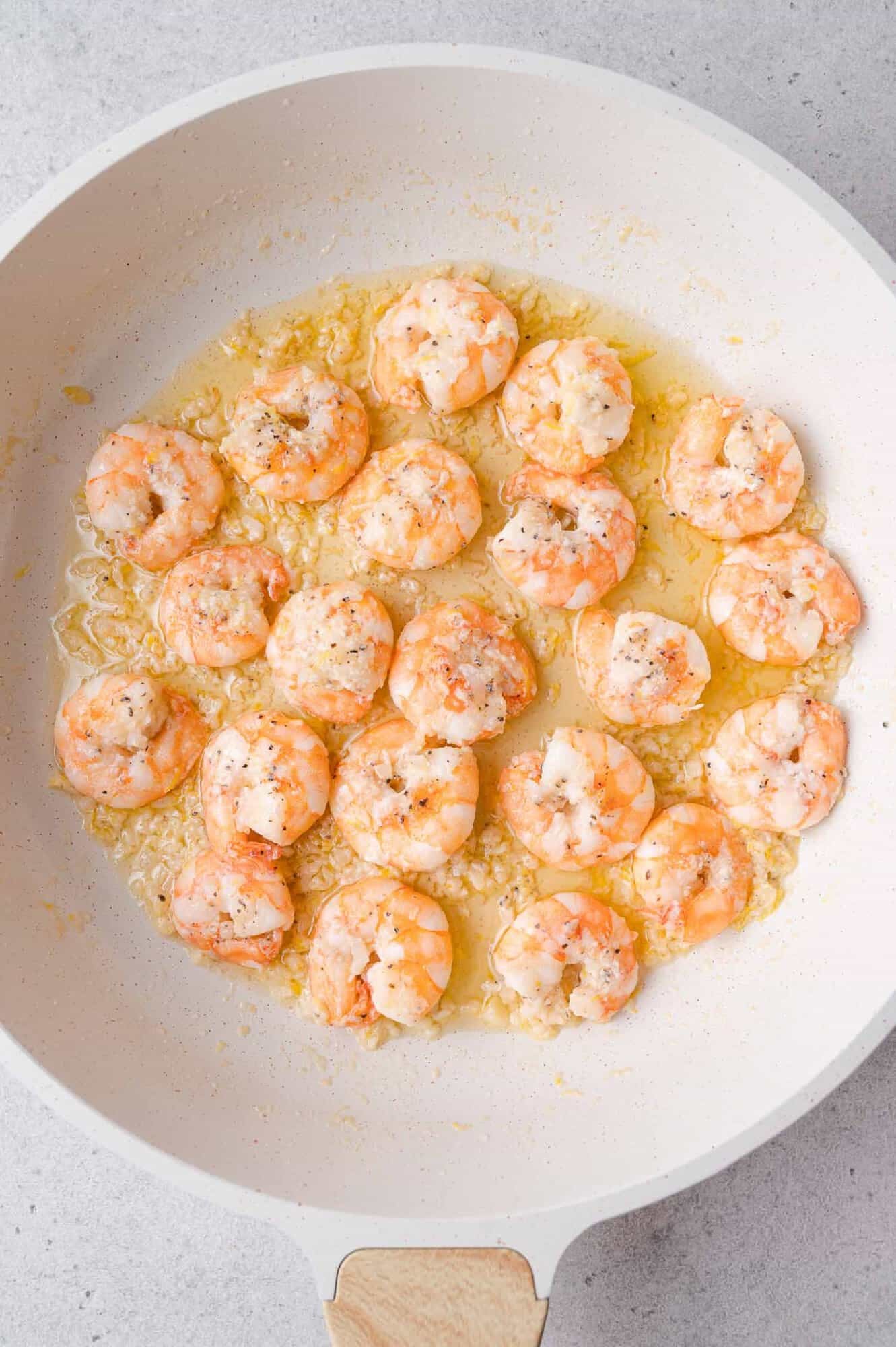 Shrimp cooked in a white skillet with minced garlic and butter, topped with black pepper and herbs on a light gray surface.