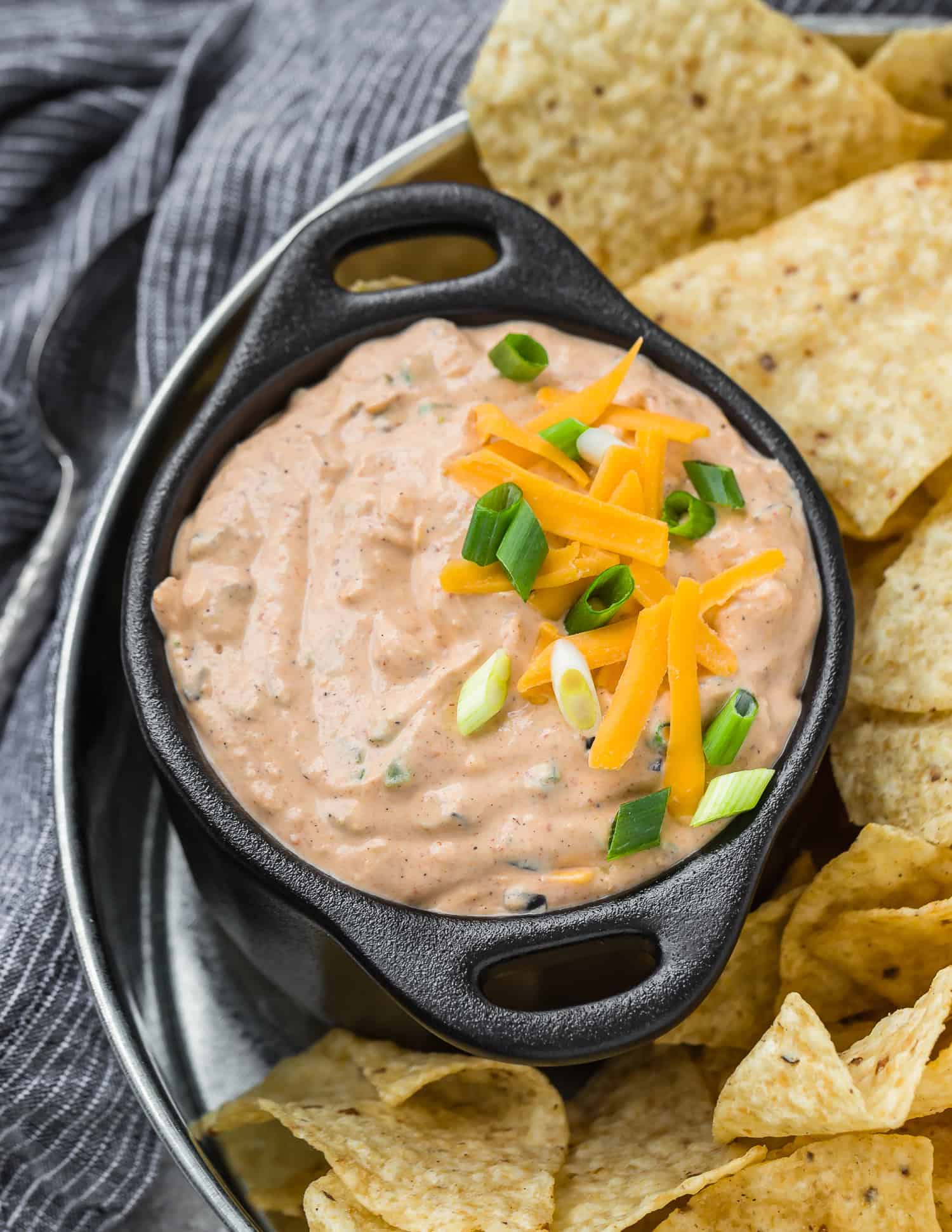 A bowl of creamy Mexicali dip topped with shredded cheddar cheese and chopped green onions, served with tortilla chips on the side.