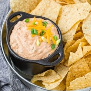 A black bowl of creamy Mexicali dip topped with shredded cheese and green onions, surrounded by tortilla chips on a metal tray.
