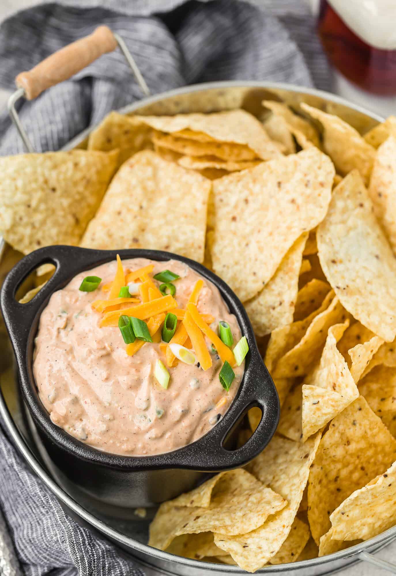 A bowl of creamy dip topped with shredded cheese and green onions is surrounded by tortilla chips on a metal tray.