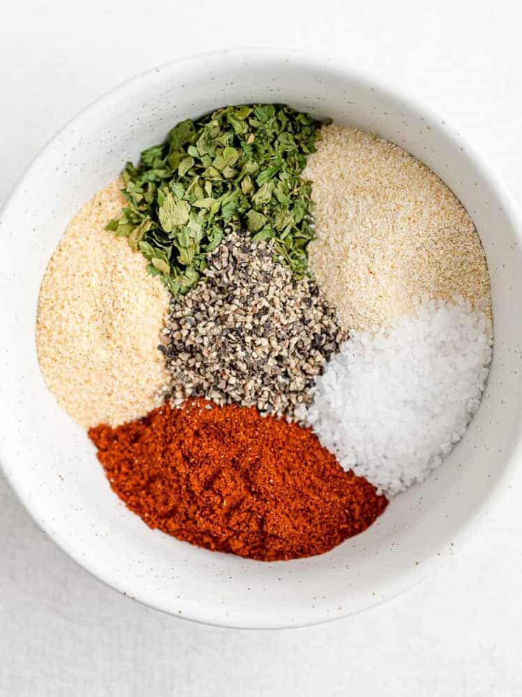A white bowl contains separate piles of dried herbs, black pepper, salt, paprika, and garlic powder on a white surface.