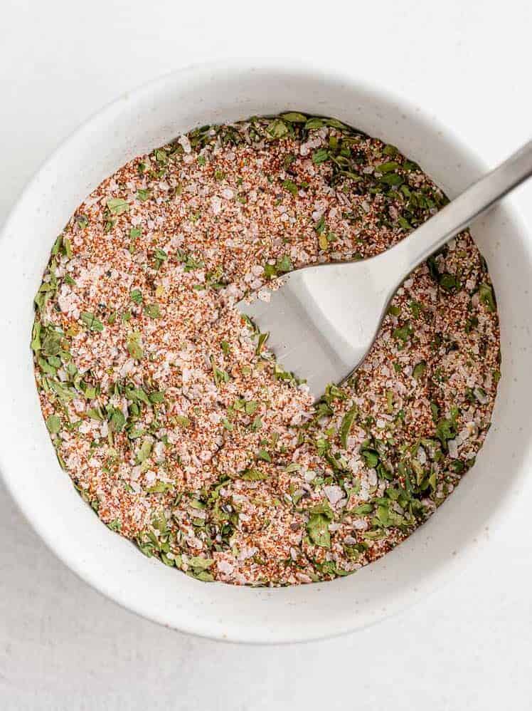 A white bowl filled with a mixed dry seasoning blend, including green herbs and red spices, with a metal fork resting inside.