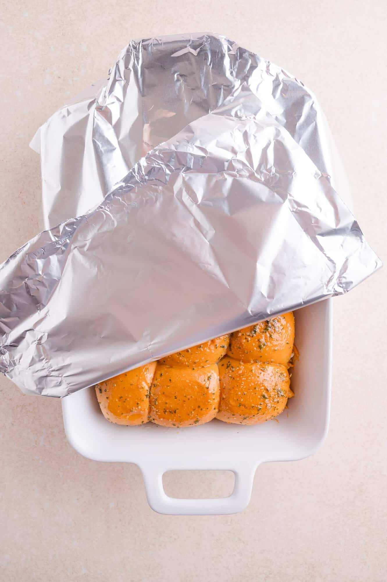 A white baking dish with six herb-topped buns partially covered by a sheet of aluminum foil, on a light-colored surface.