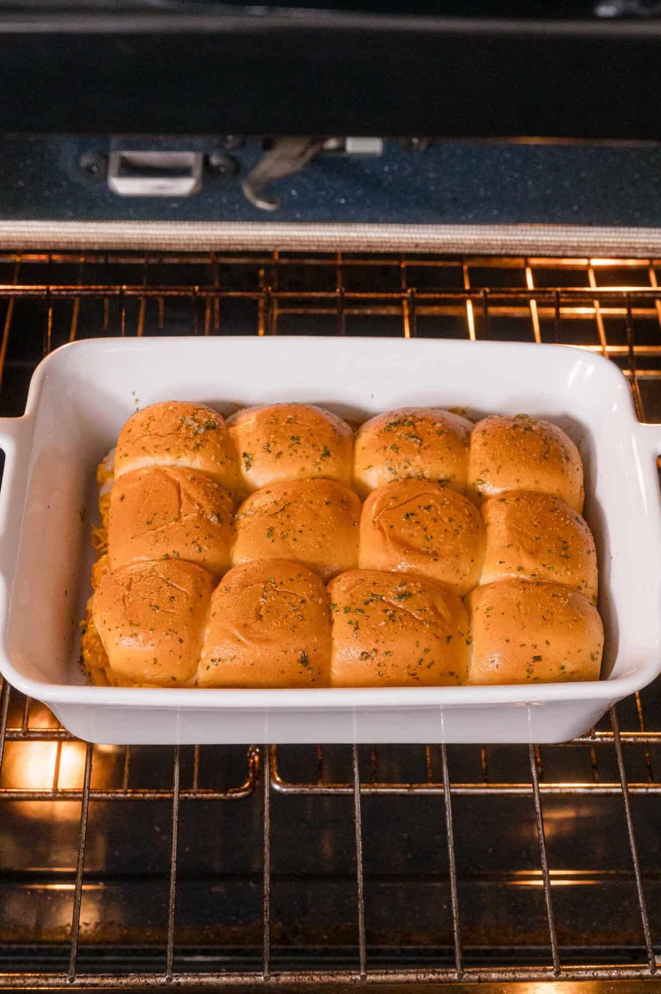 A white baking dish with twelve golden brown slider rolls sprinkled with herbs is on the oven rack, baking.