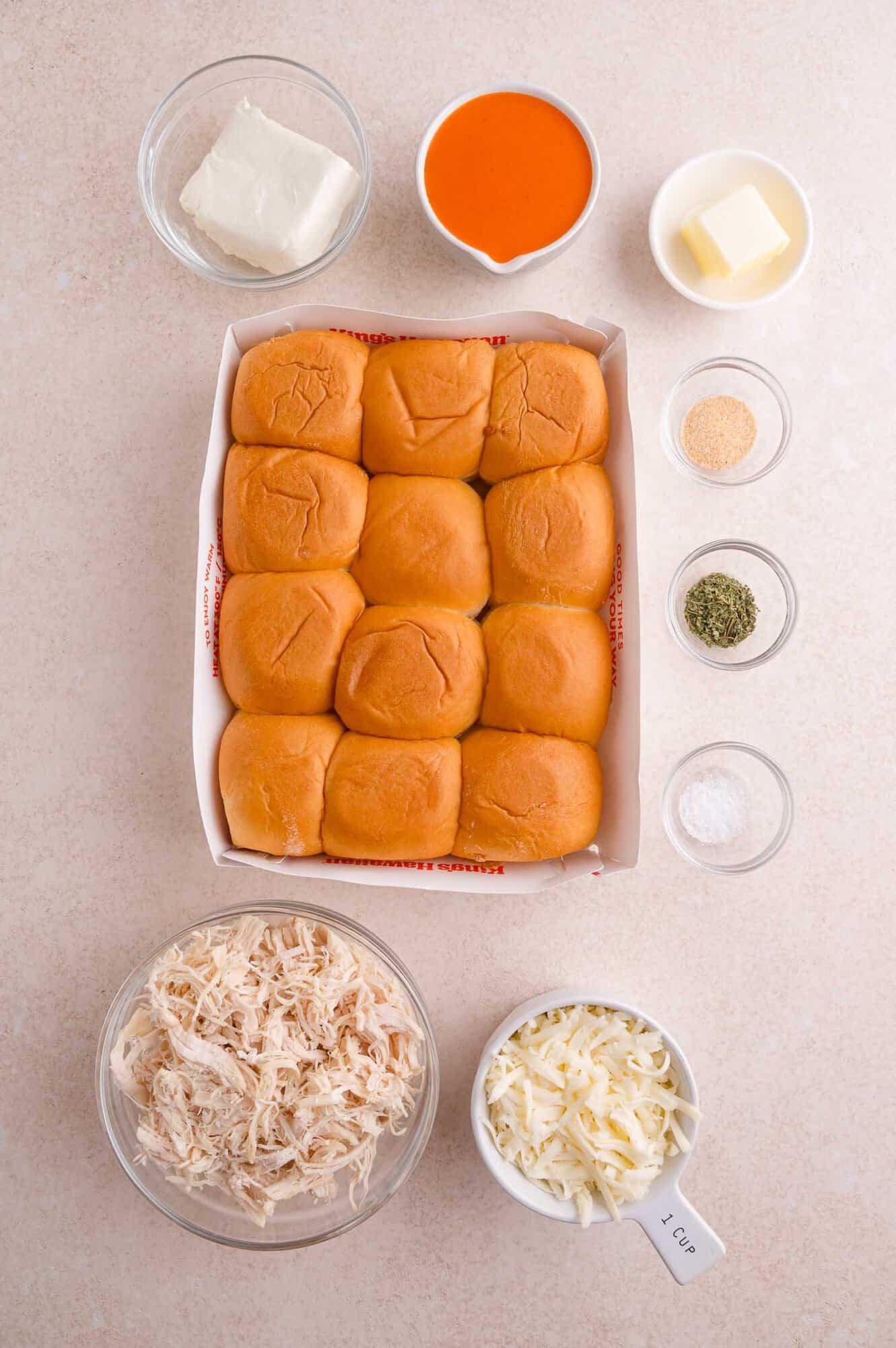 Twelve slider rolls, shredded chicken, shredded cheese, cream cheese, hot sauce, butter, and small bowls of spices arranged on a light surface.