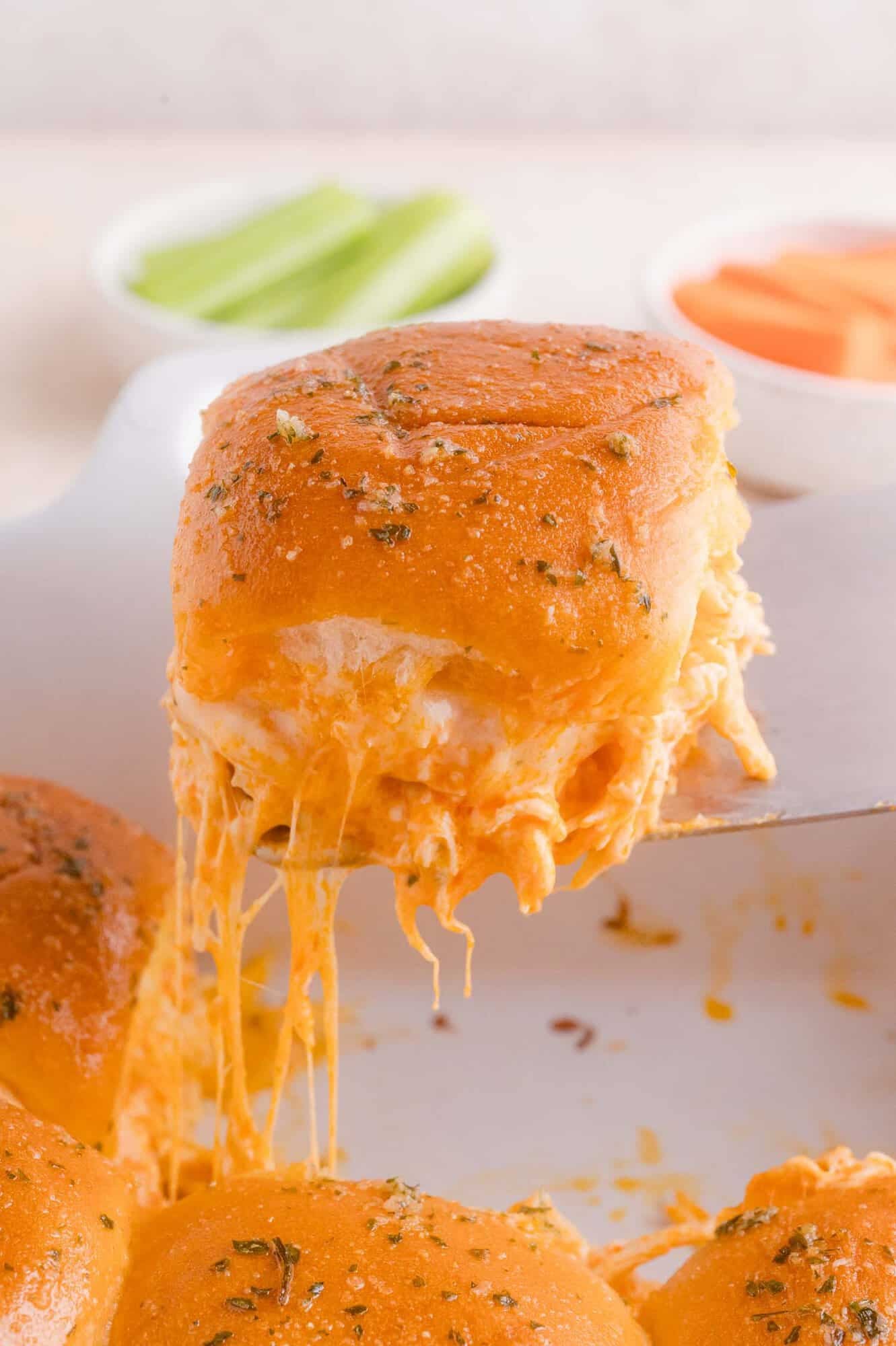 A cheesy buffalo chicken slider with herbs is lifted from a tray, showing melted cheese. Blurry bowls with celery and carrot sticks are visible in the background.