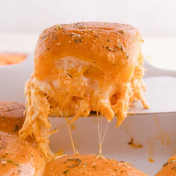 A close-up of a cheesy Buffalo chicken slider being lifted from a pan with melted cheese stretching from the bun.