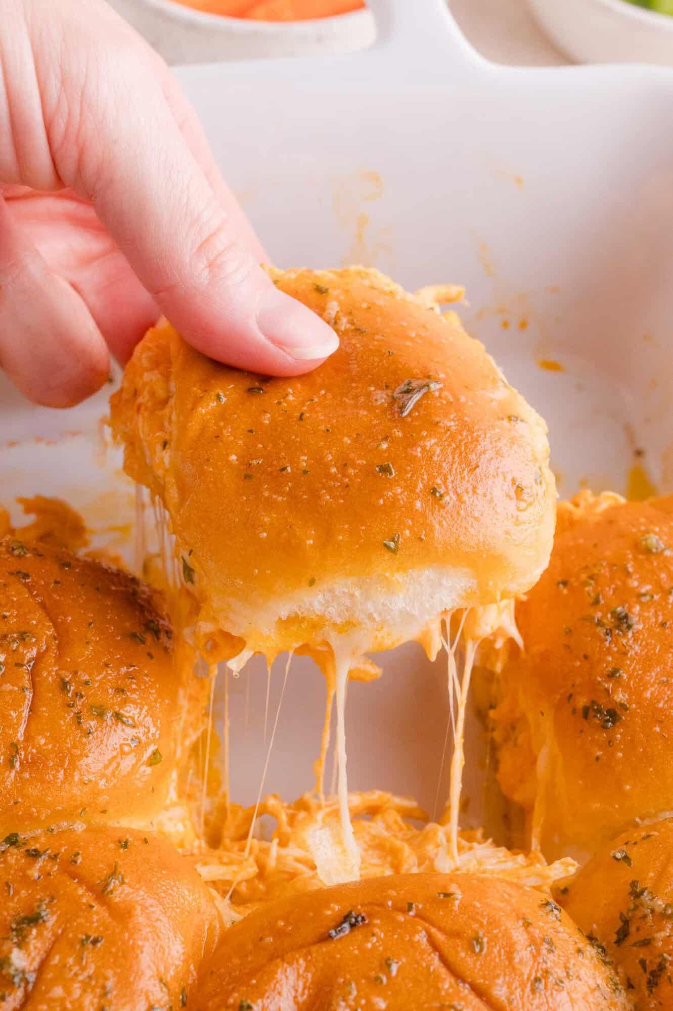 A hand lifts a cheesy, golden-brown slider bun from a baking dish, with melted cheese stretching between the rolls.