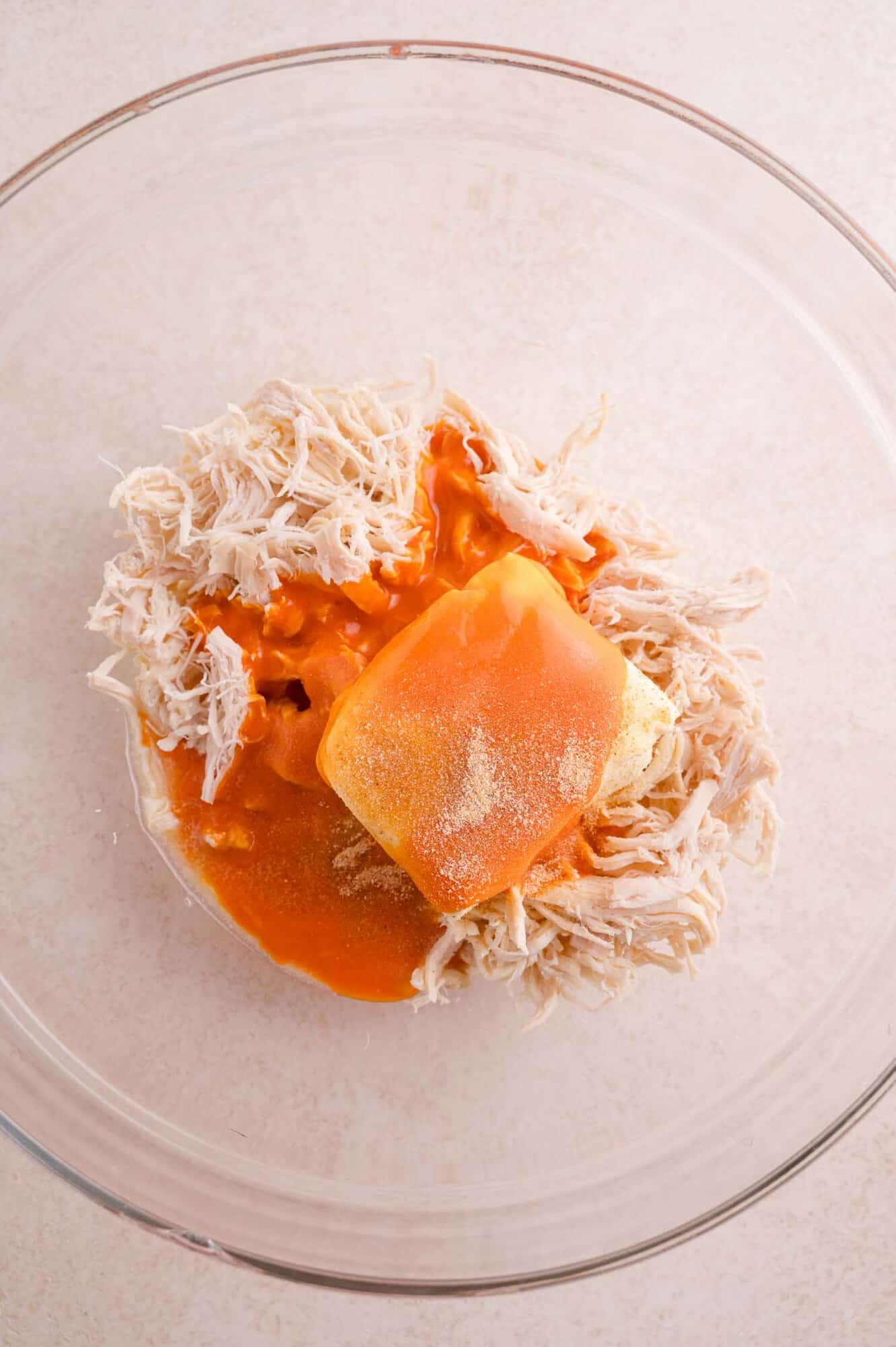 A glass bowl containing shredded chicken, buffalo sauce, a block of butter, and seasoning on a light surface.
