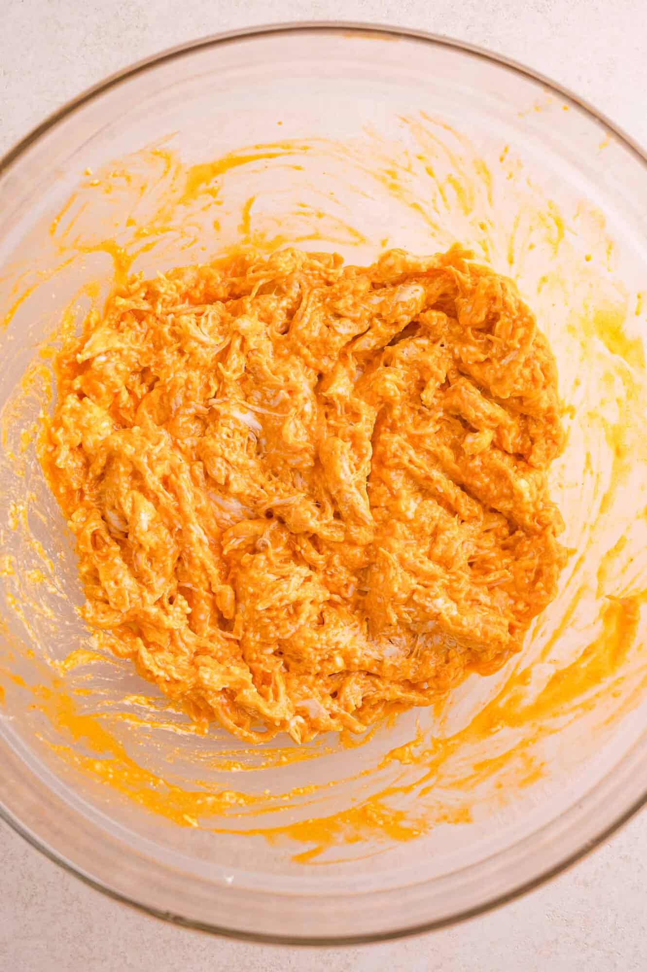 A glass bowl containing a mixture of shredded cheese and hot sauce, partially blended together, on a light countertop.