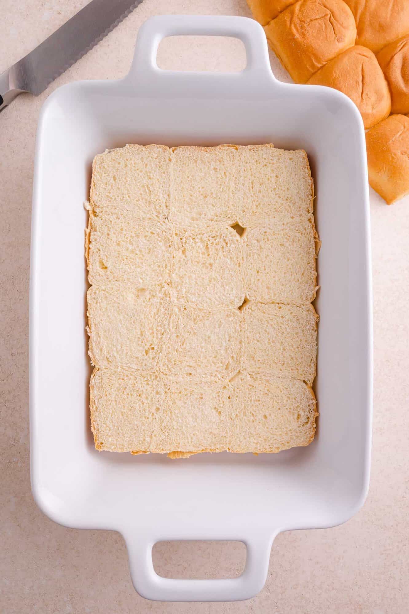 A white baking dish with a layer of sliced bread rolls arranged neatly; a bread knife and more rolls are visible nearby.