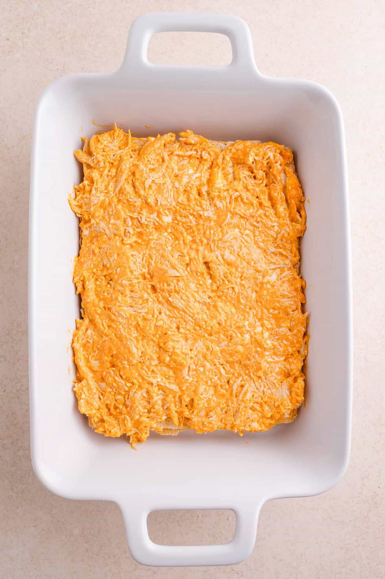 Rectangular white baking dish with a layer of chicken, hot sauce, and cream cheese mixture, evenly spread on the bottom, photographed from above on a light countertop.