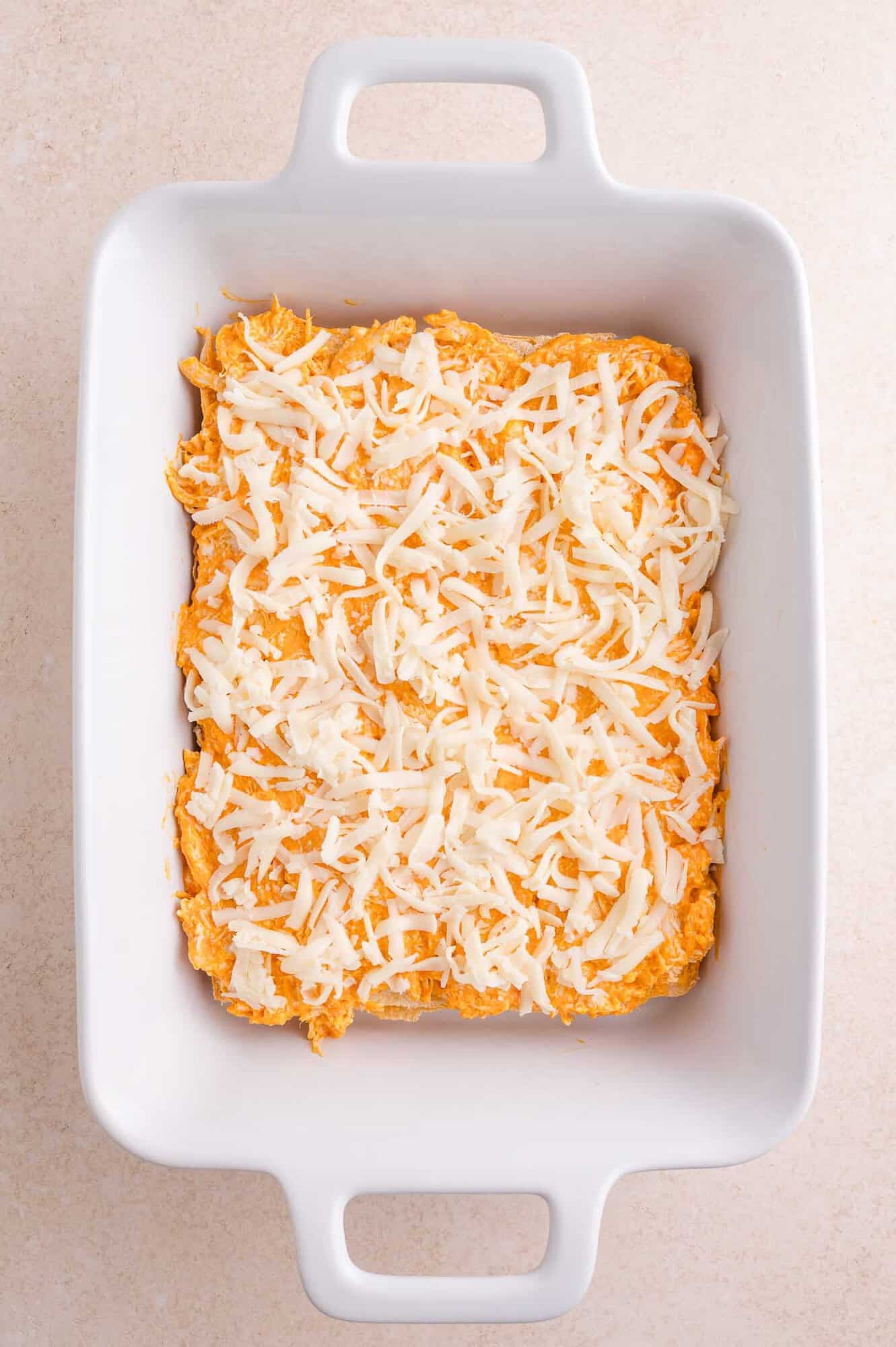 Rectangular white baking dish with an buffalo chicken mixture topped with shredded white cheese, ready to be baked, on a light-colored surface.