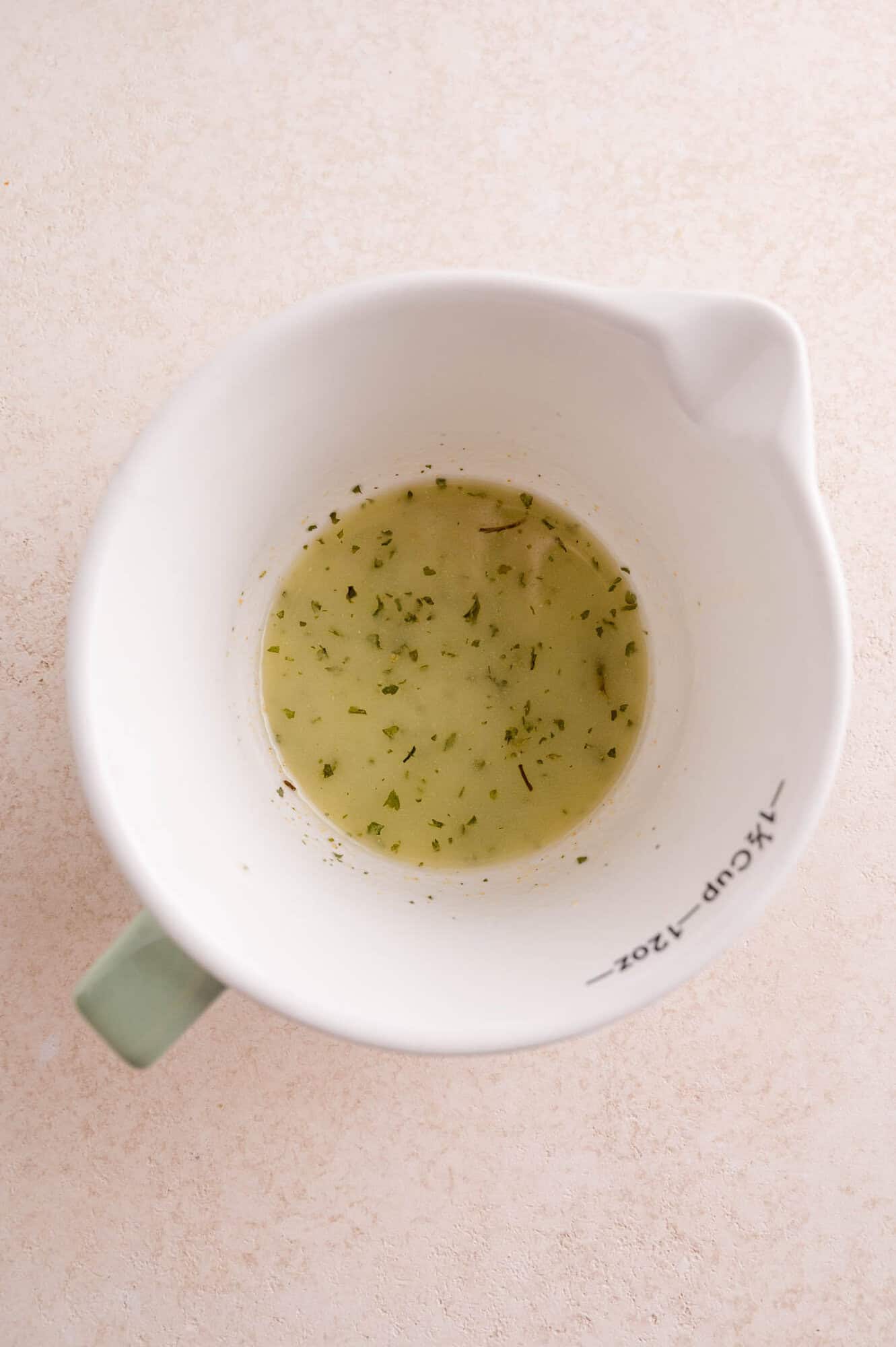 A white measuring cup with a green handle contains a small amount of seasoned butter with herbs, sitting on a light-colored surface.