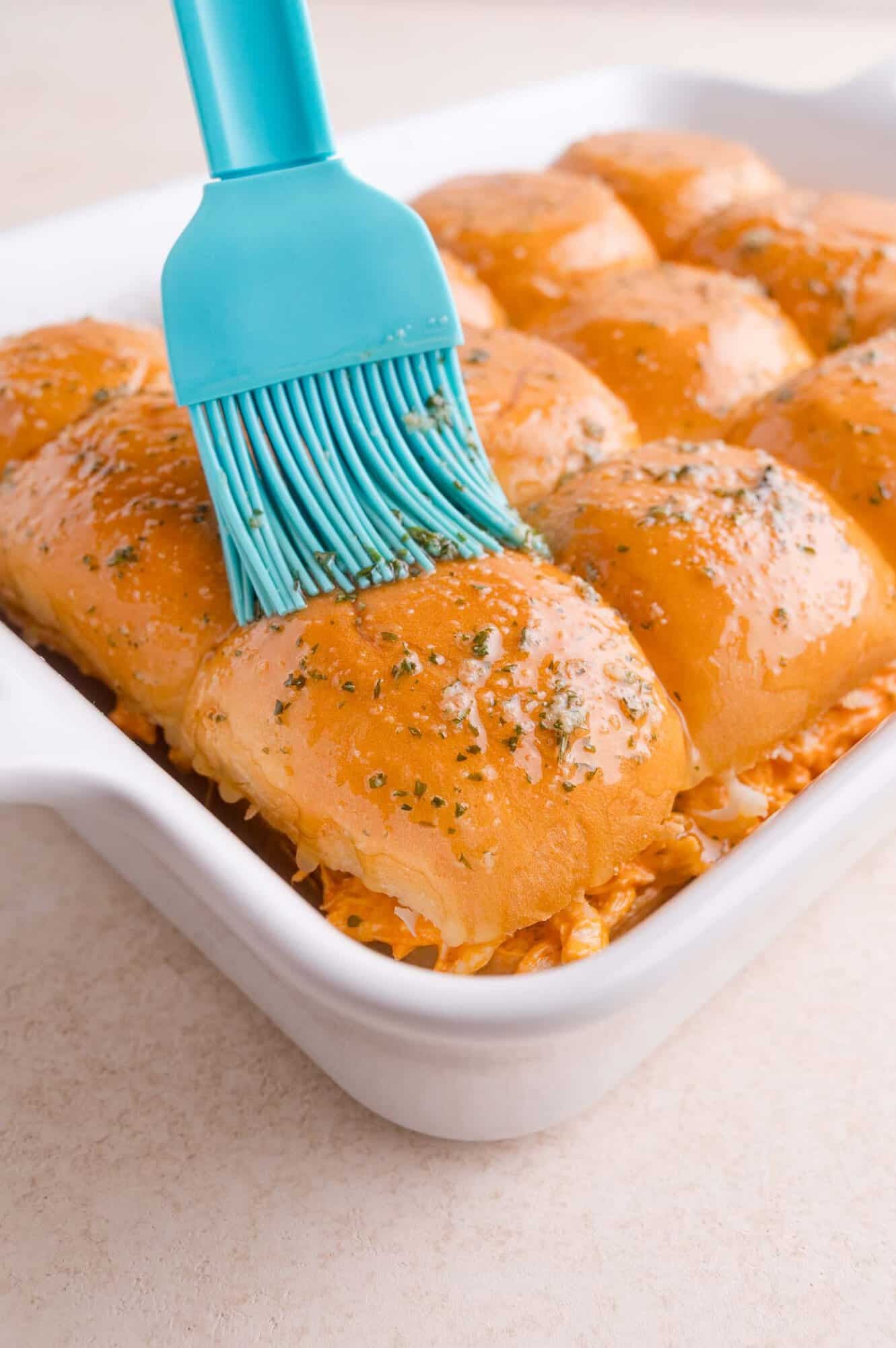 A blue brush applies melted butter to golden baked slider sandwiches topped with herbs in a white baking dish.