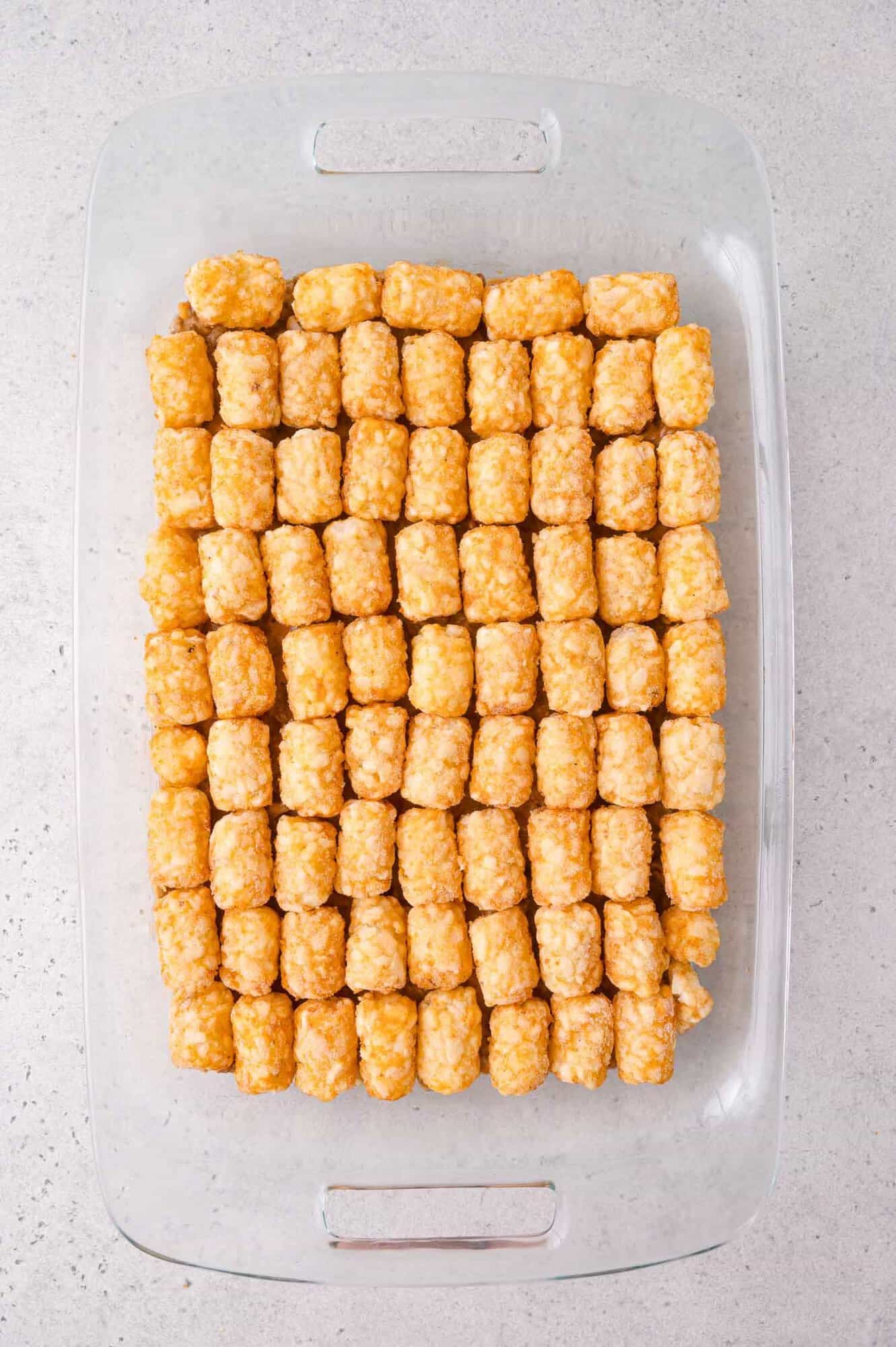 A glass baking dish filled with neatly arranged rows of golden tater tots on a light-colored countertop.